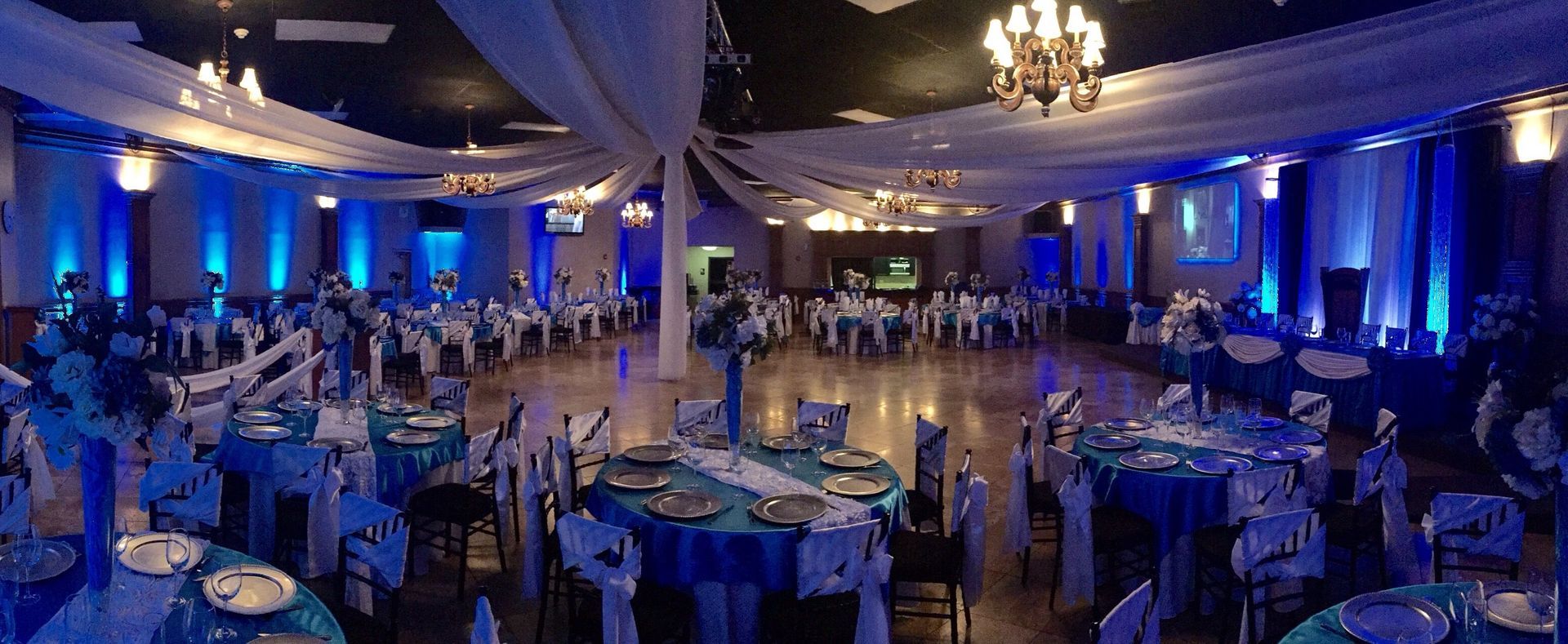 Blue-lit banquet hall with round tables and chairs set for an event under chandeliers.