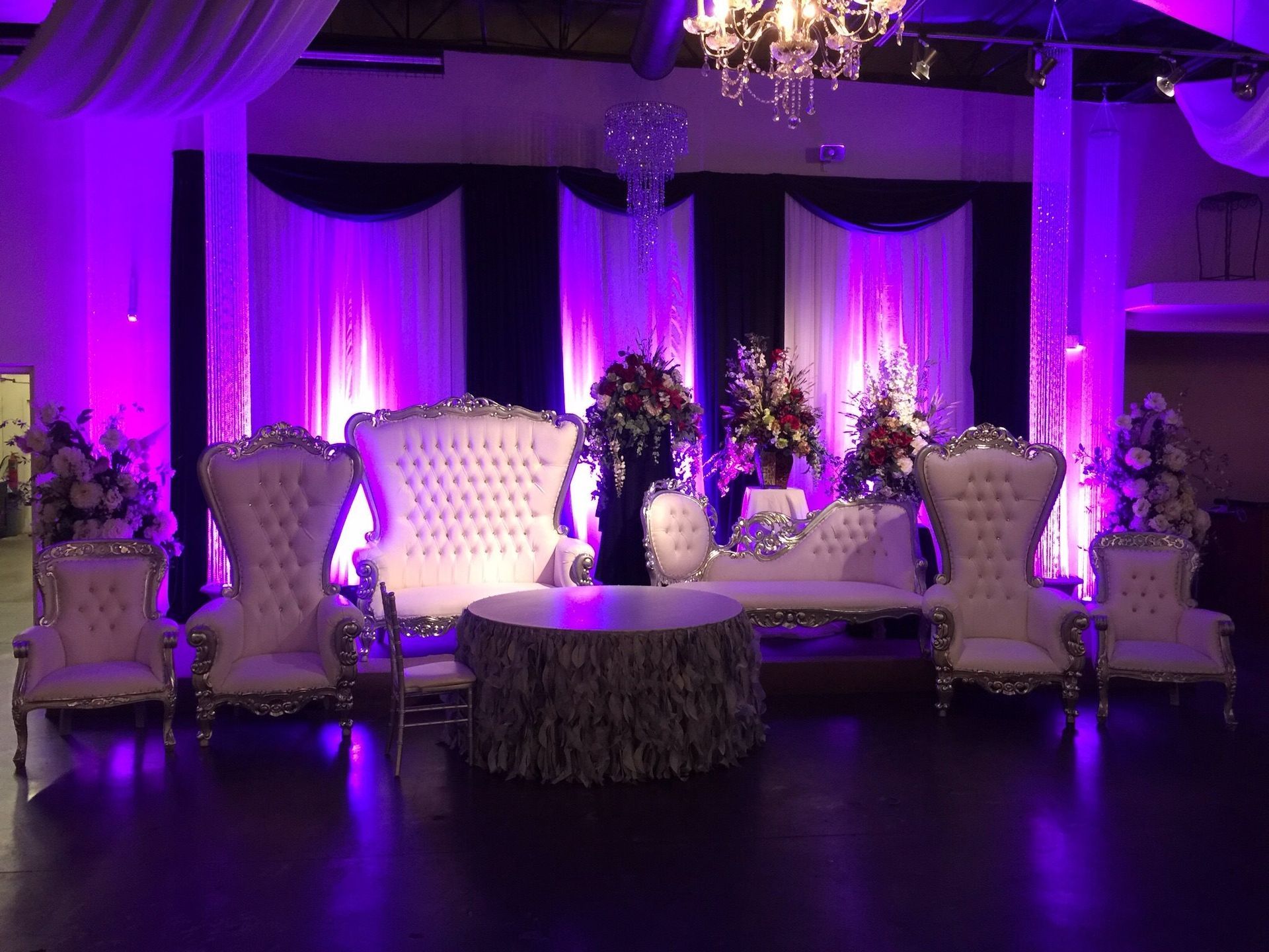 Elegant purple-lit event lounge with white tufted sofas, chairs, and floral arrangements on a draped stage