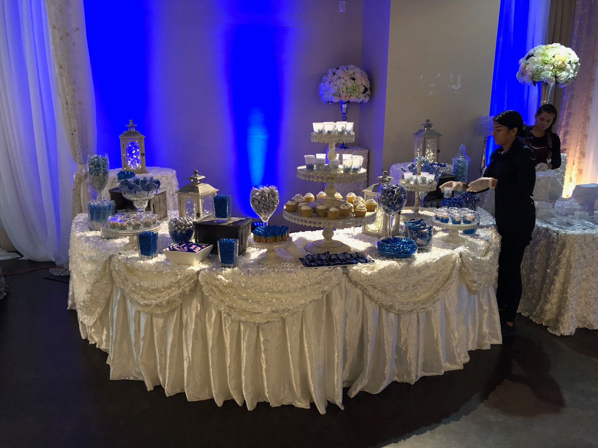 Blue-lit banquet hall with round tables set for a formal event and a chandelier overhead