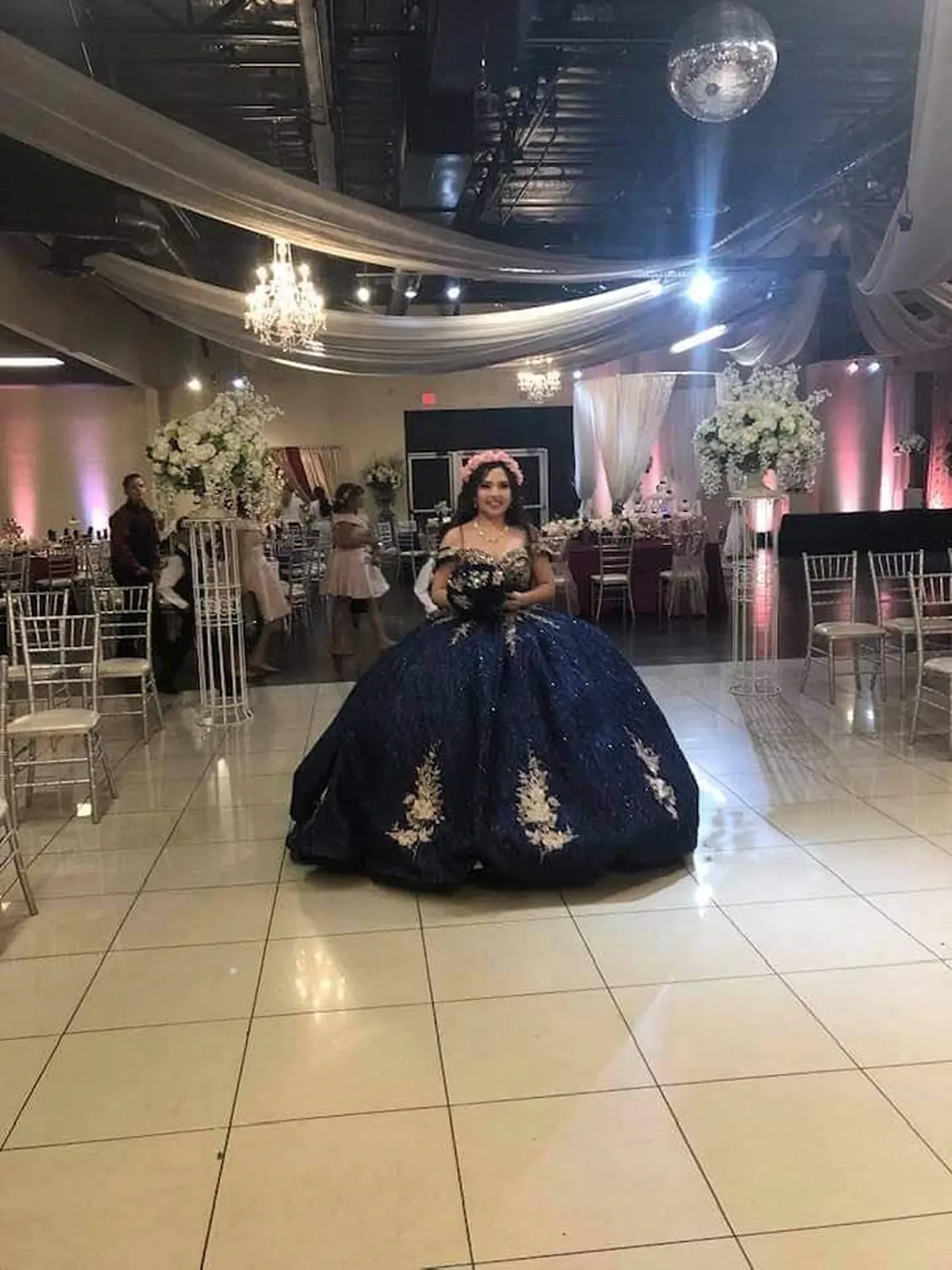 Person in a dark blue ball gown standing in a decorated ballroom under hanging drapes and lights.