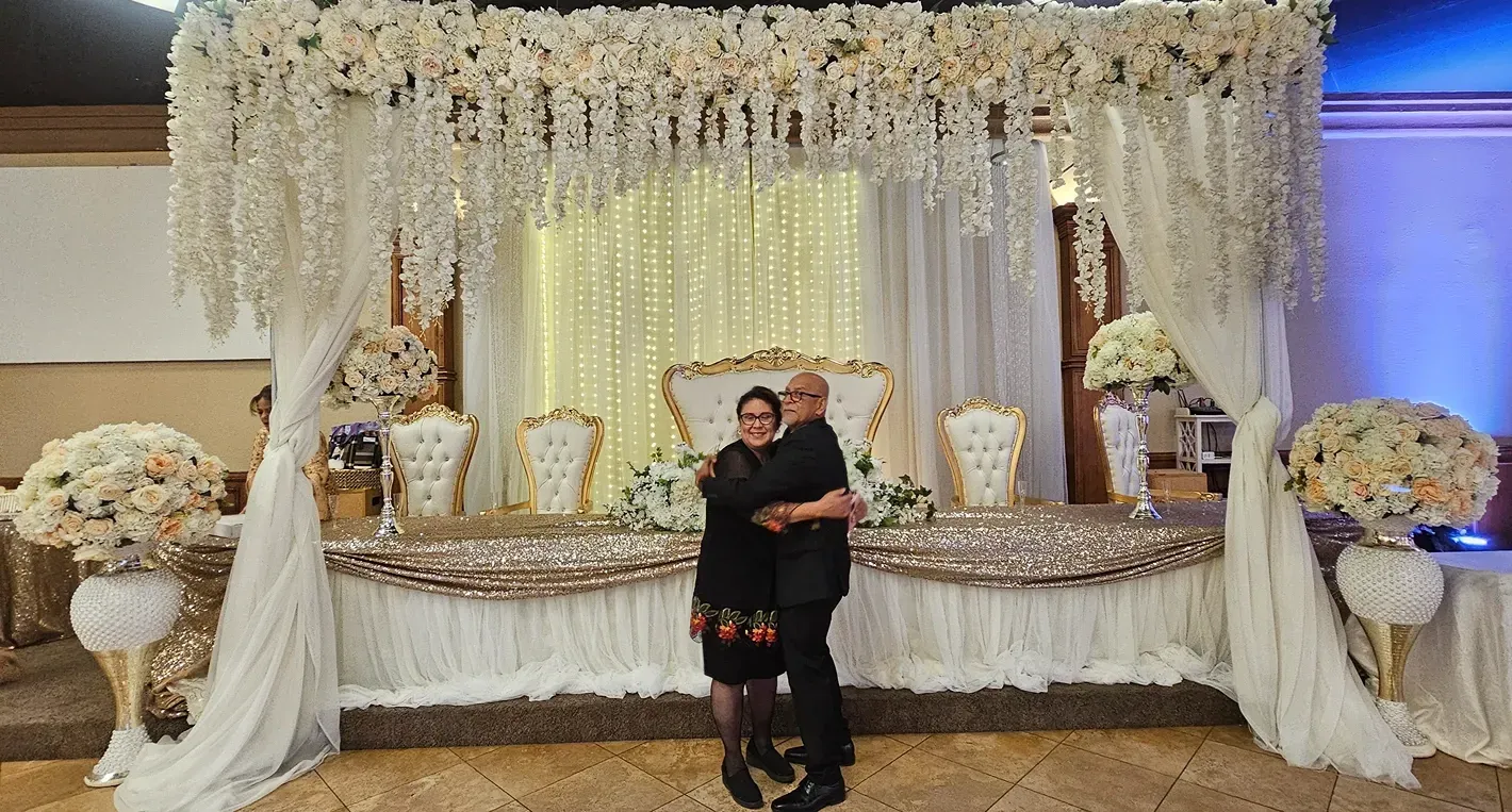 Couple posing in front of a white floral wedding stage with gold accents and draped lights