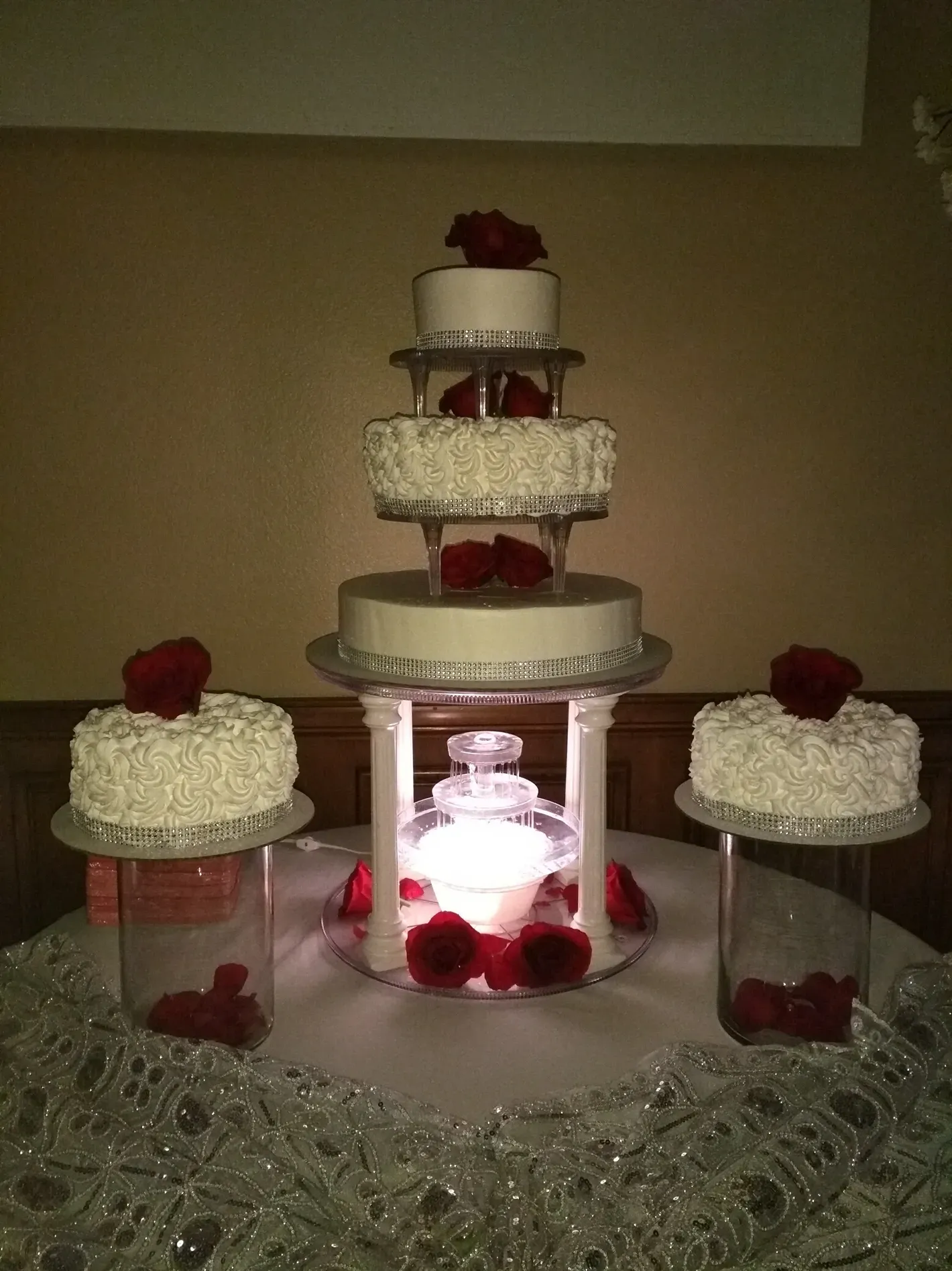 Tiered white wedding cake with red roses, displayed on a mirrored stand with smaller matching cakes around it.