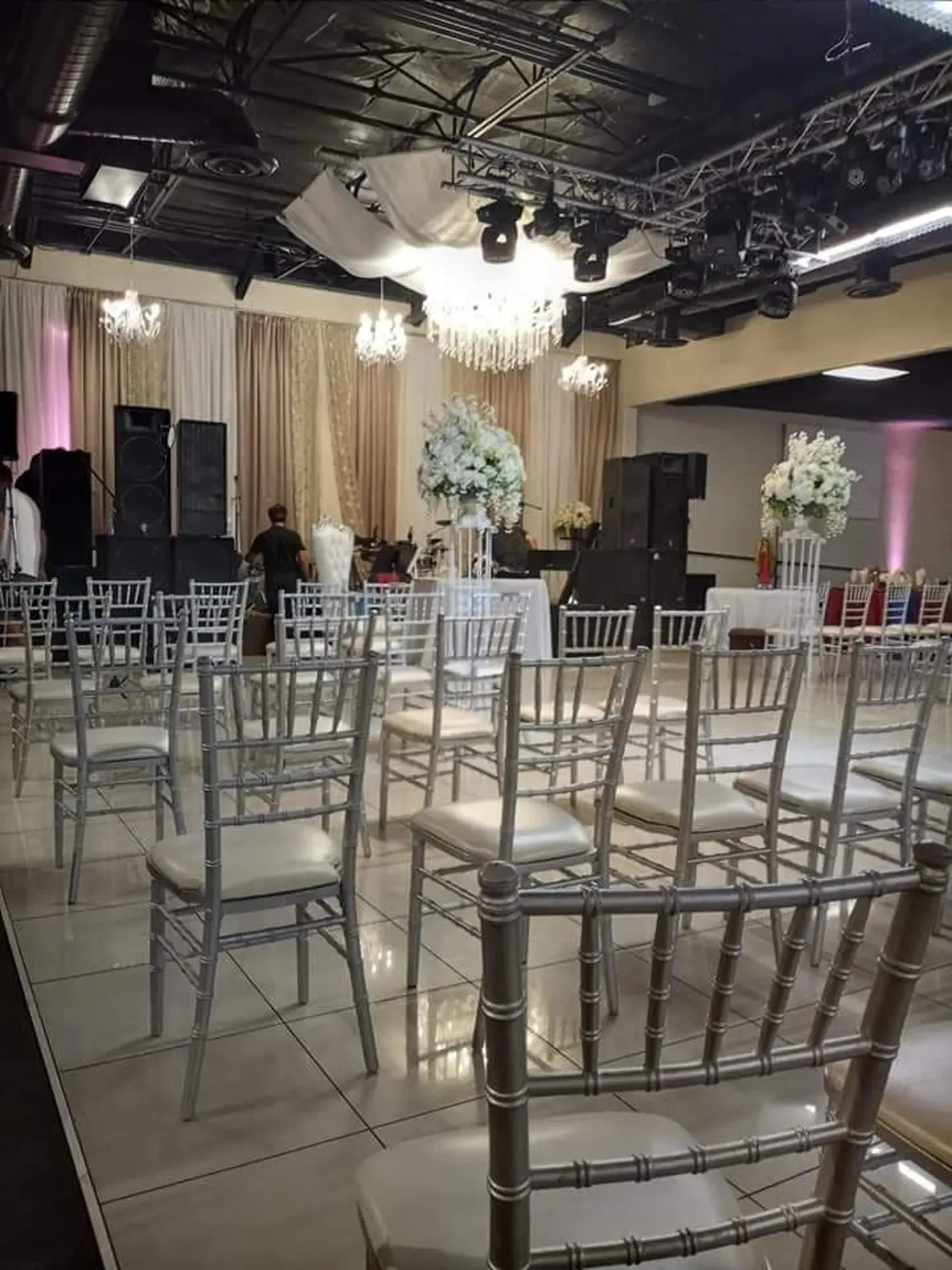 Empty banquet hall with silver chairs, round tables, and a decorated stage under ceiling lights