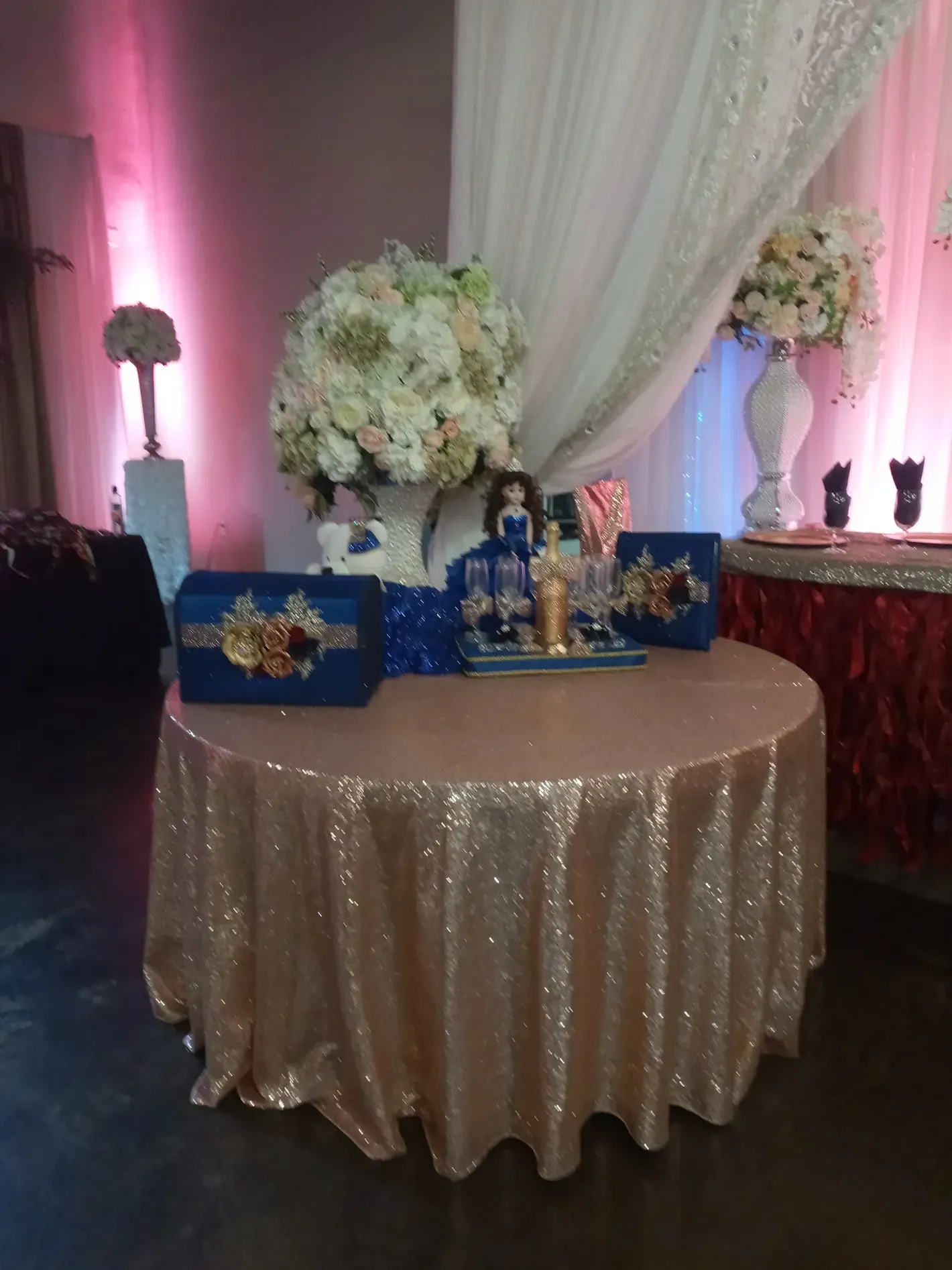 Decorated dessert table with gold sequin cloth, blue accents, floral arrangements, and pink uplighting
