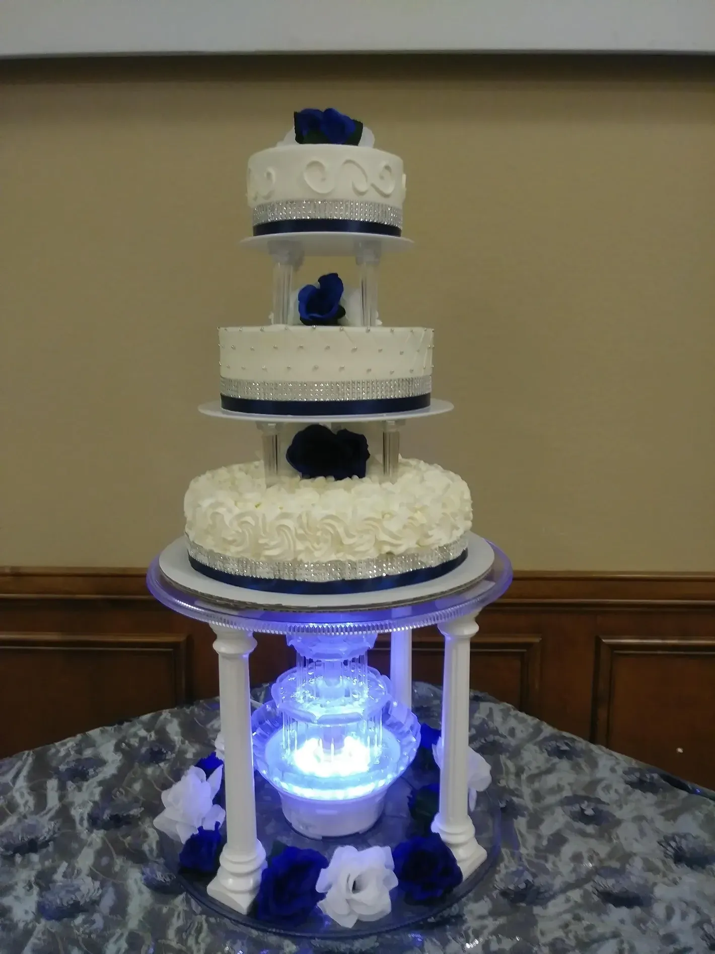 Three-tier white cake with blue accents on a lit pedestal display stand in a room