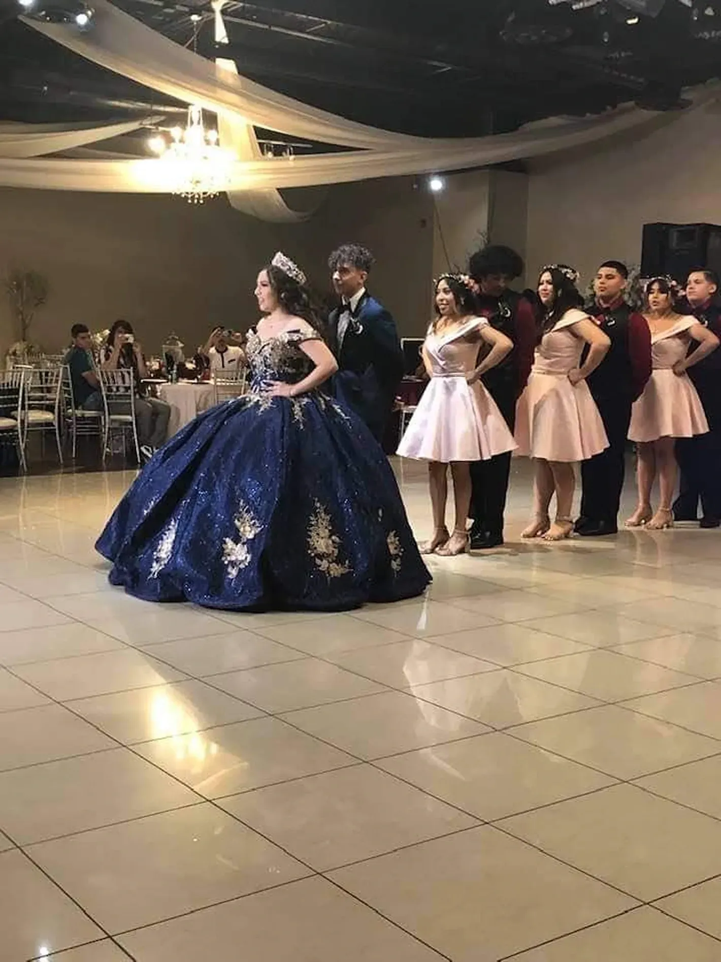 Young woman in a blue ball gown stands beside girls in pink dresses at a formal dance floor.