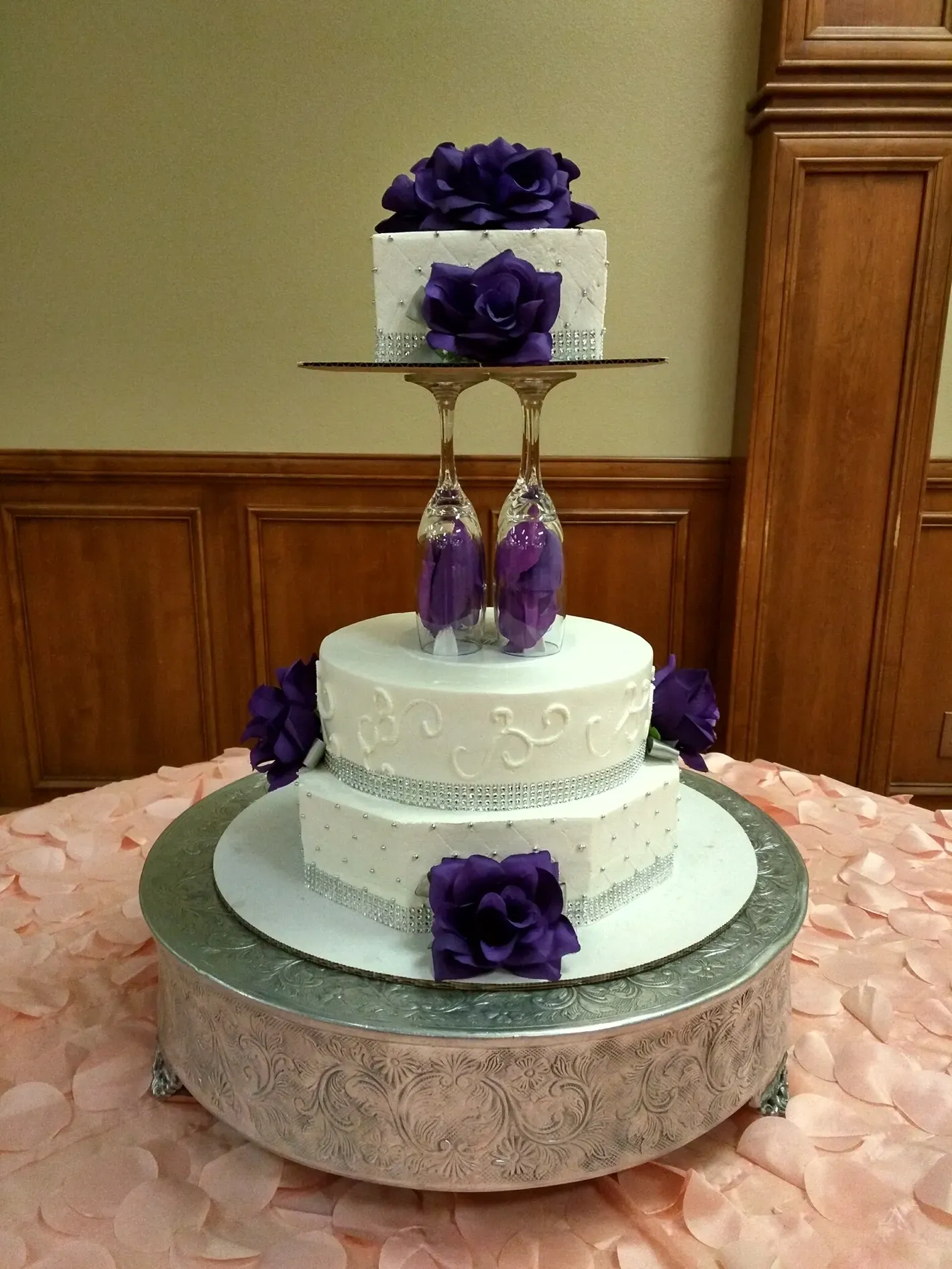 Three-tier white cake with purple flowers on a silver stand, set on a round table.