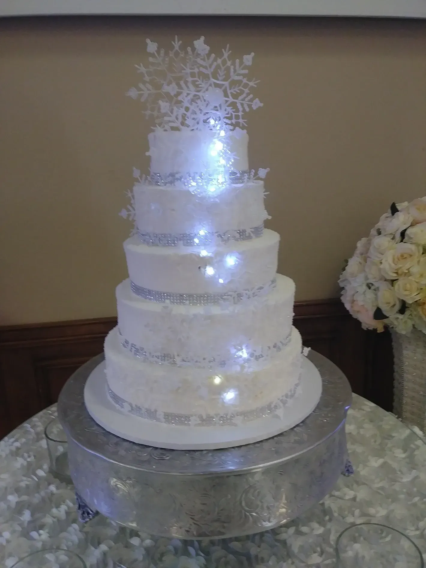 Five-tier white wedding cake with silver accents and sparkling lights on a silver pedestal