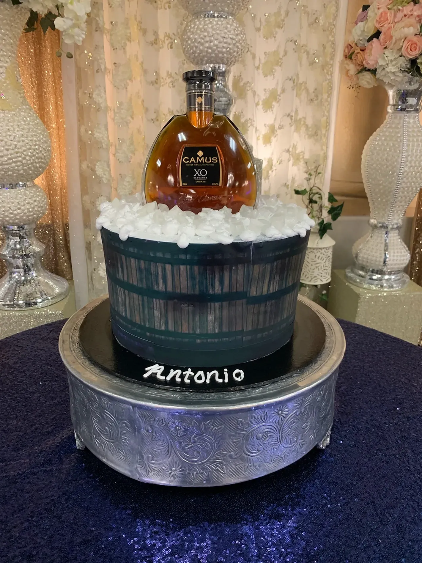 Decorative whiskey barrel cake with bottle topper and the name “Antonio” on a silver pedestal table