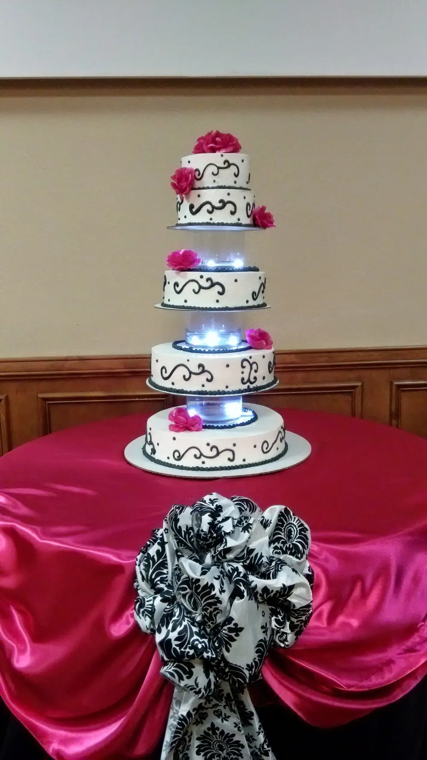 Four-tier white cake with pink flowers on a red satin-draped table, with a black-and-white bow in front.