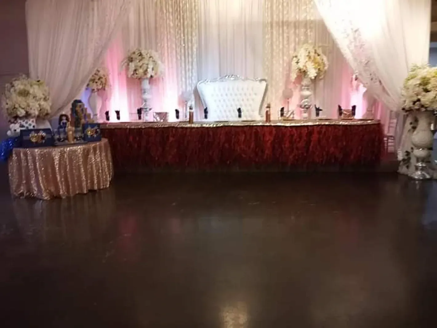 Wedding reception hall with decorated head table, white drapes, floral arrangements, and dark dance floor