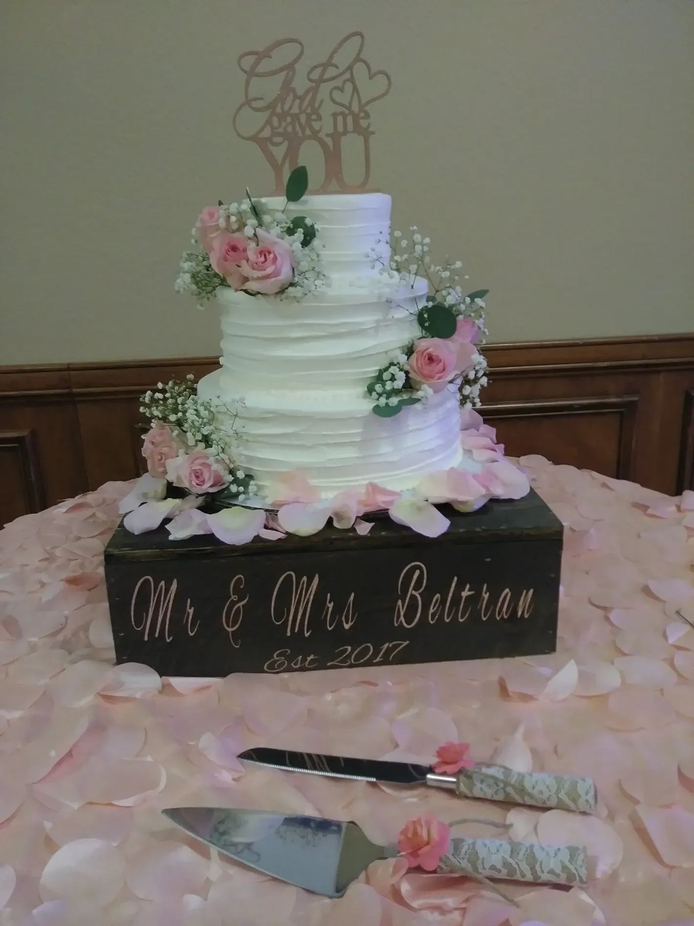 White tiered wedding cake with pink flowers and a wooden topper on a decorated table.