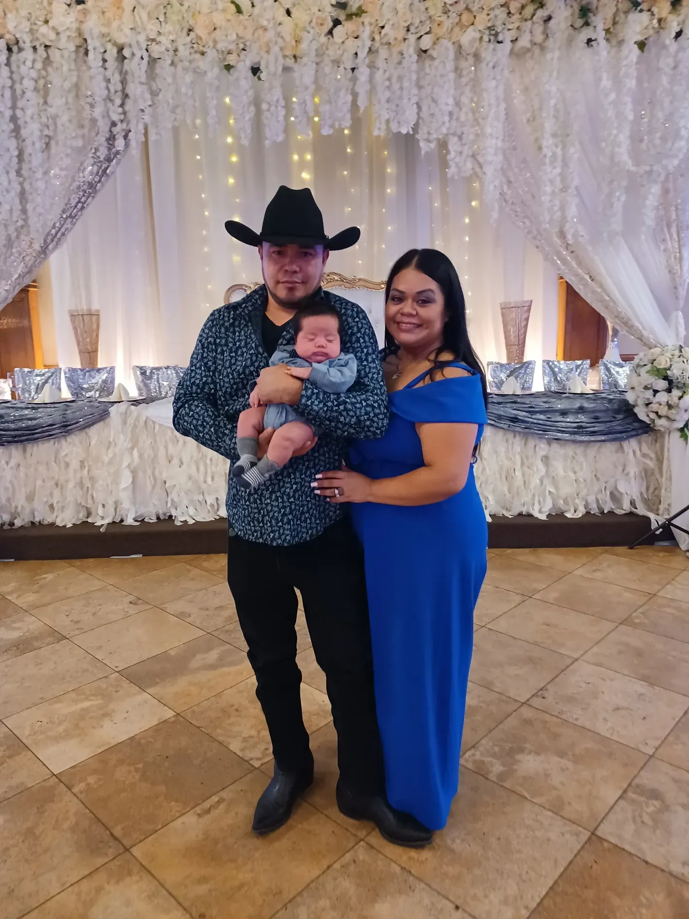 Man in cowboy hat and woman in blue dress holding a baby at a decorated event backdrop