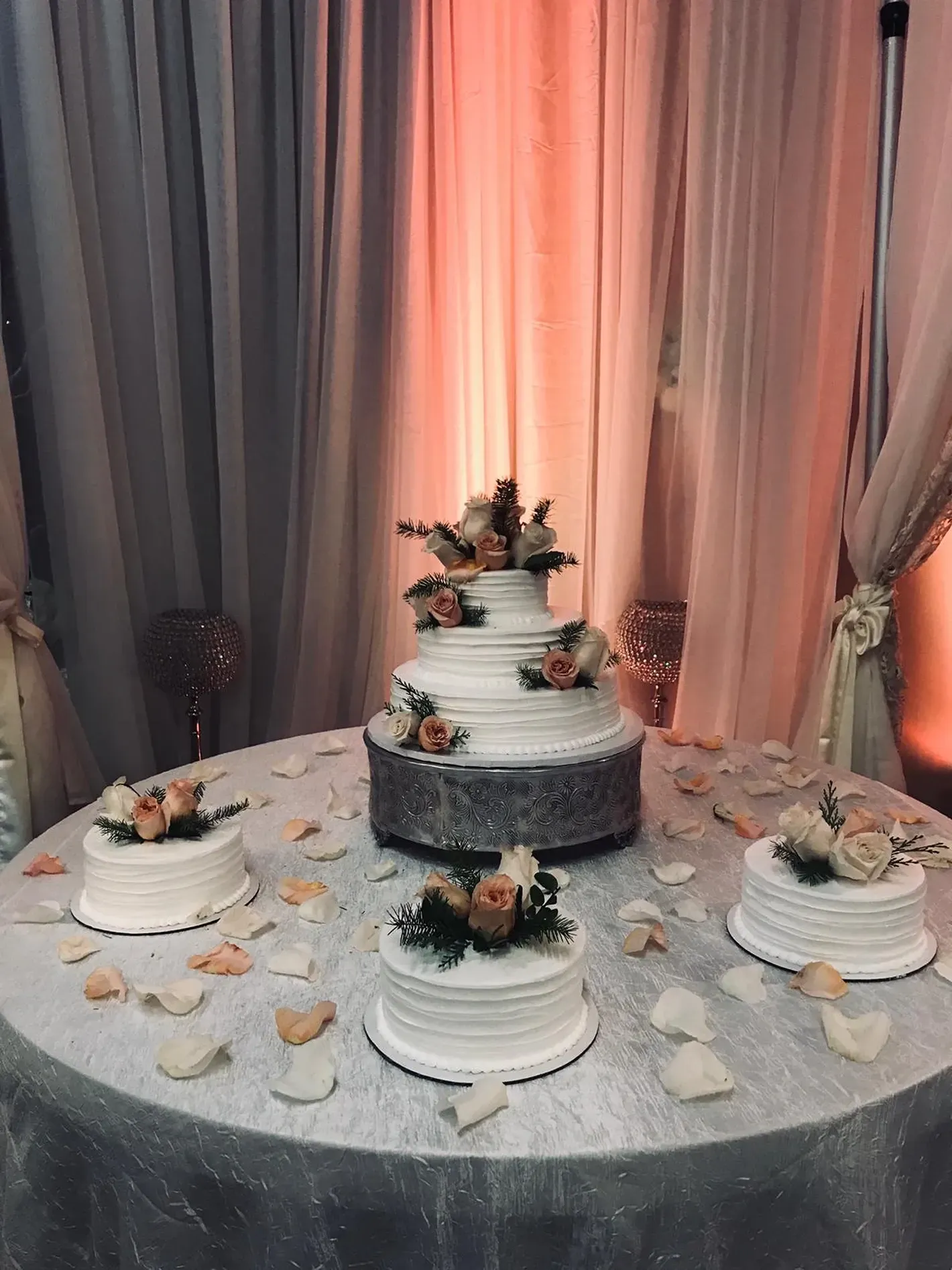 Wedding cake display on a silver table with a tiered white cake, two small cakes, and white petals