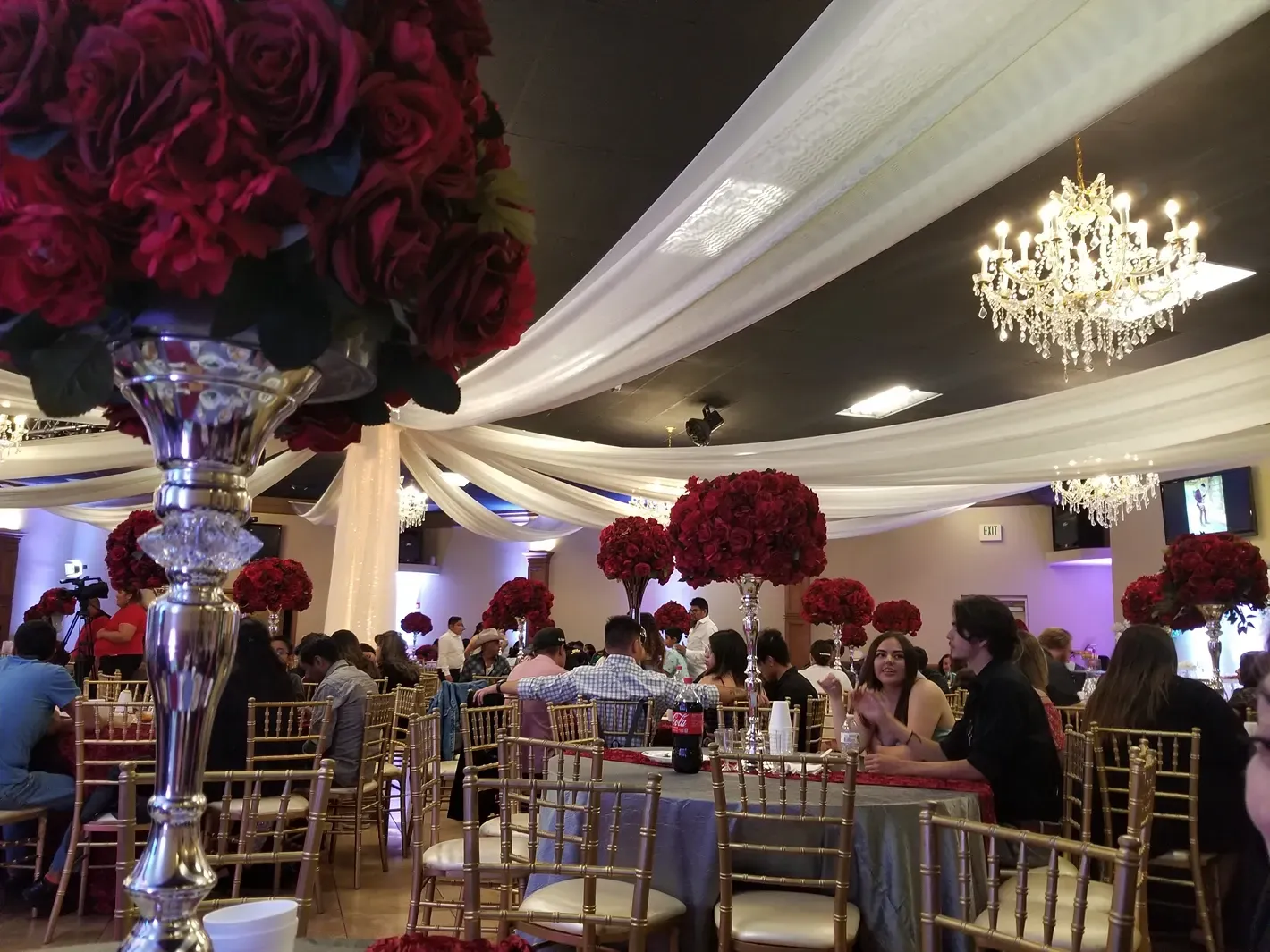 Elegant banquet hall with gold chairs, chandeliers, and red floral centerpieces under white draped ceilings