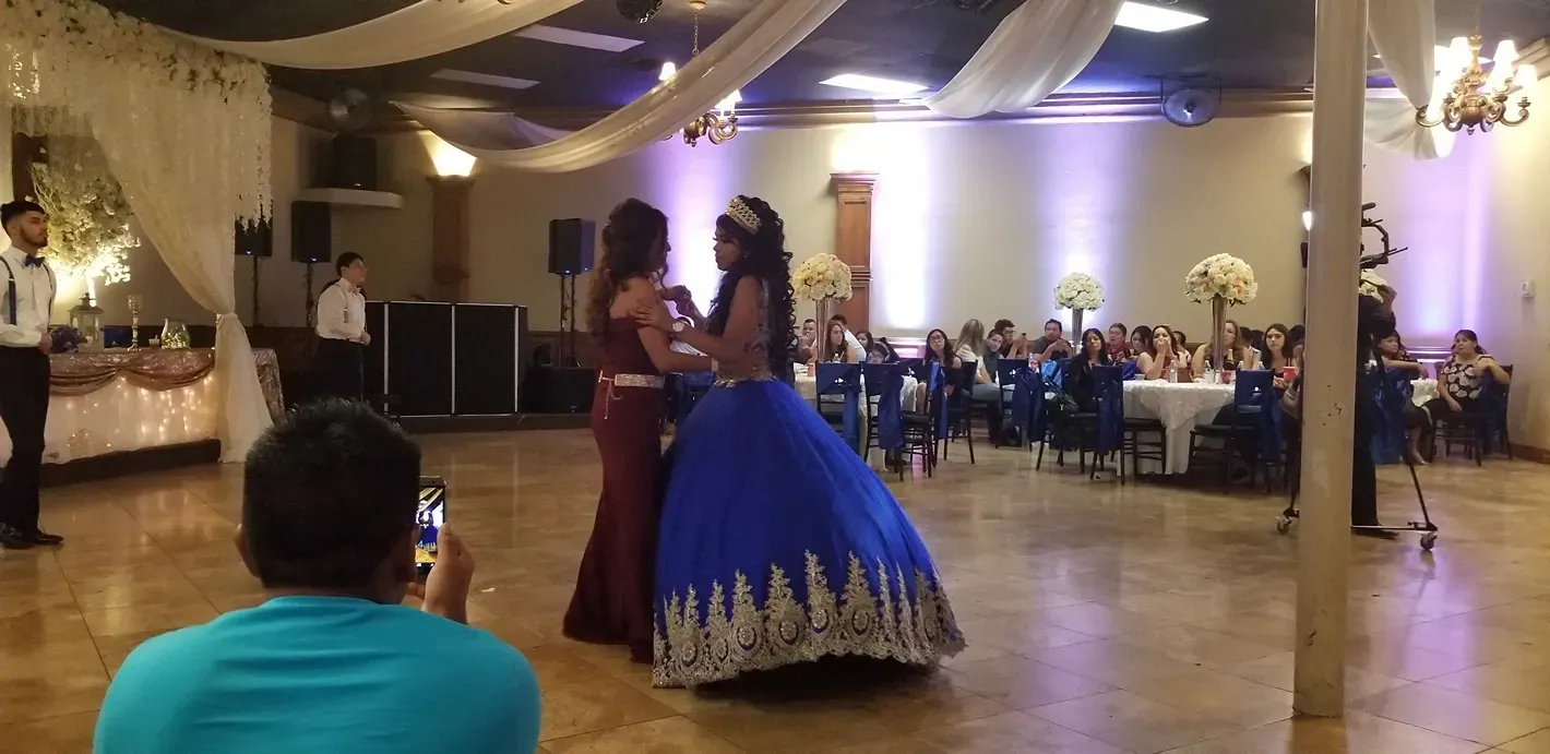 Two women in formal dresses dancing at a banquet hall reception with seated guests in the background