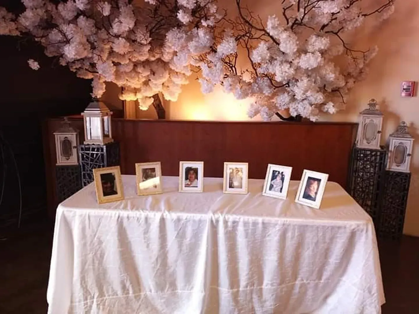 Memorial table with framed photos and white tablecloth beneath a floral backdrop and lanterns