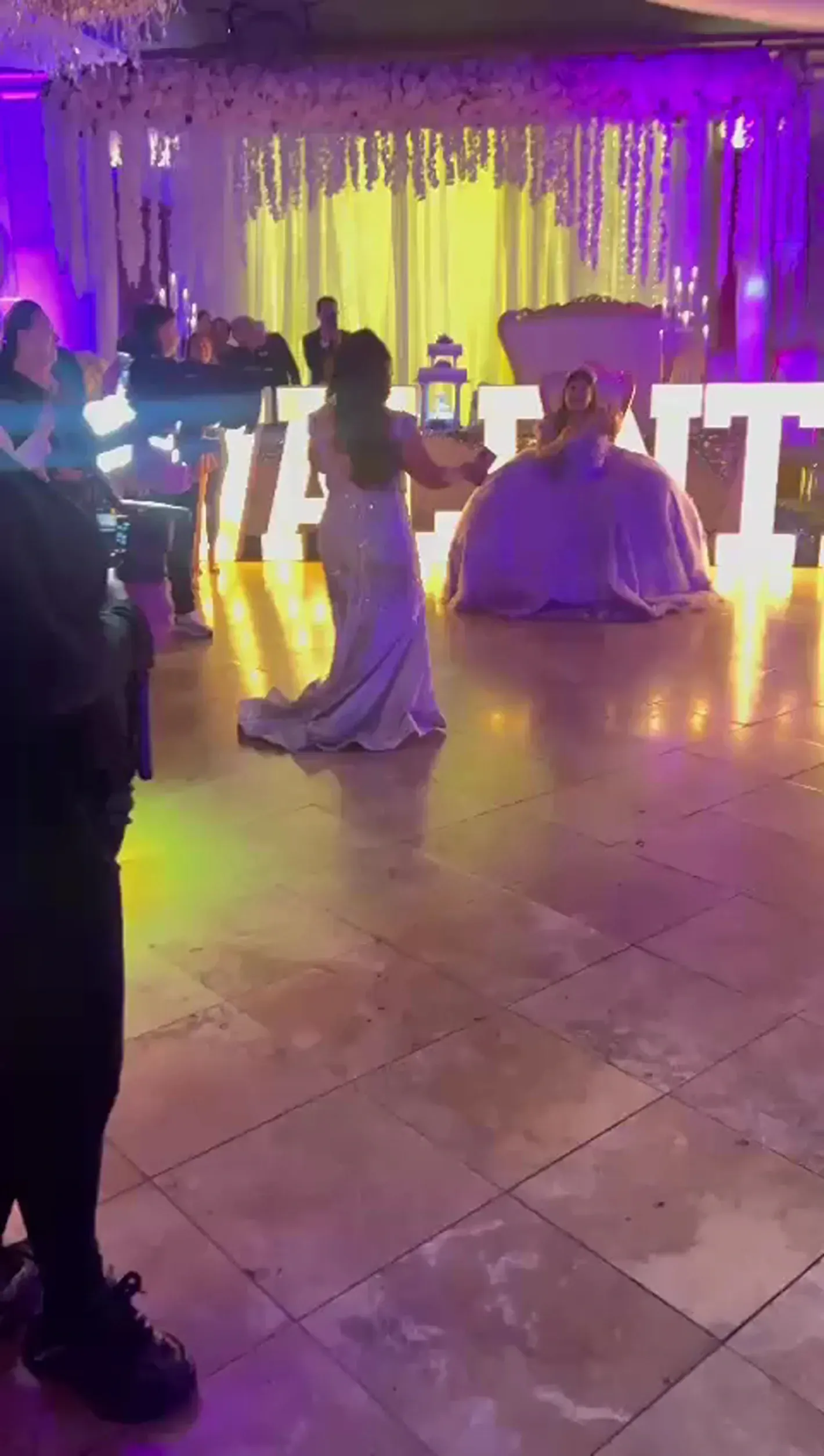 Purple-lit event hall with white drapery, glowing letters, and seated guests on a tiled floor.