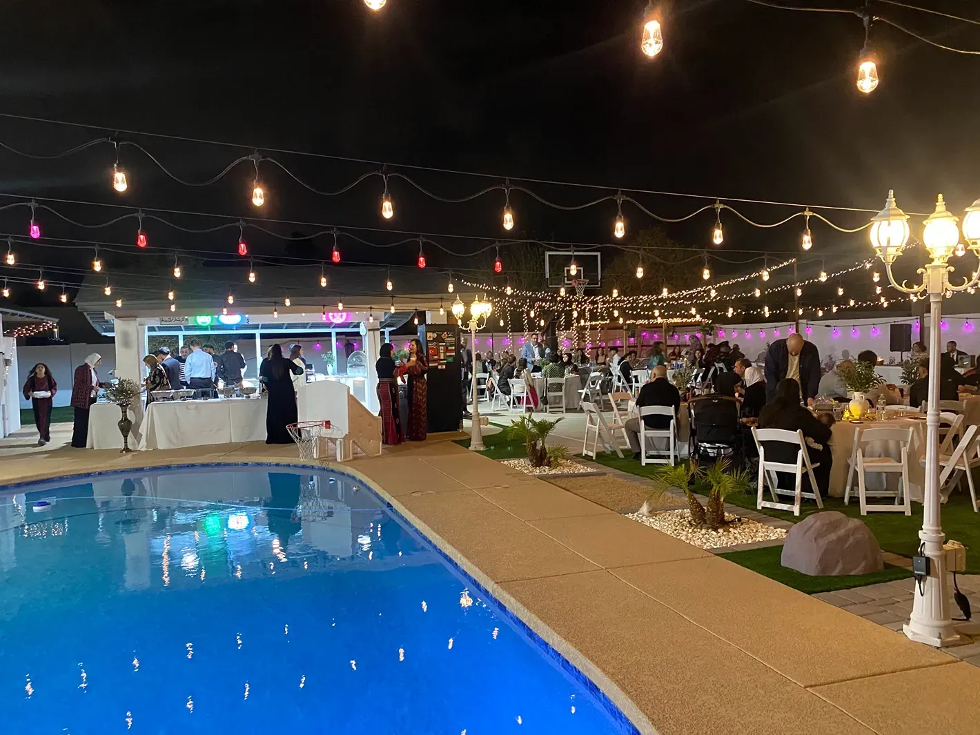 Nighttime poolside event with string lights, seated guests, and a lit blue pool near a white canopy tent