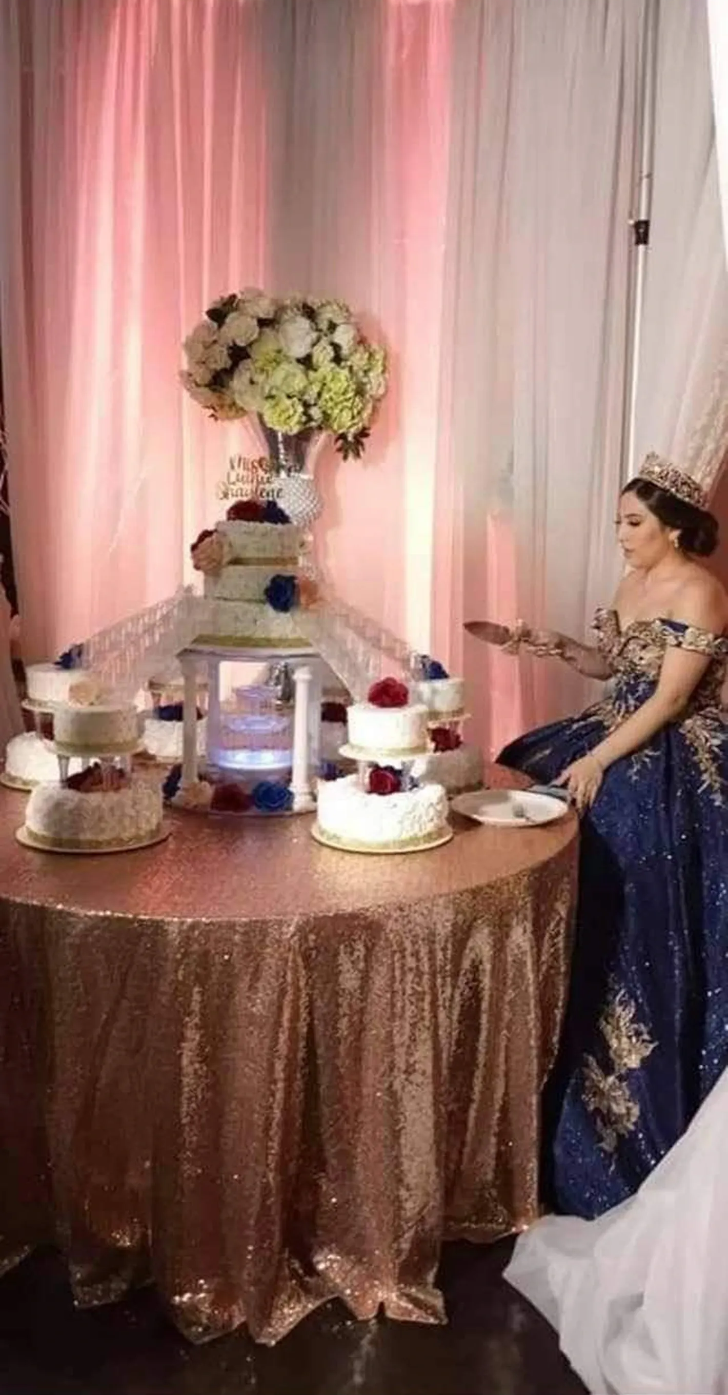 Decorated cake table with pink drapes, floral centerpiece, and a woman in a blue gown seated nearby