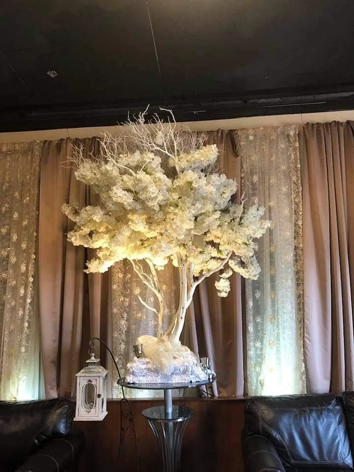 Decorative white flowering tree centerpiece in a living room, lit by warm uplights, with curtains and leather sofas.