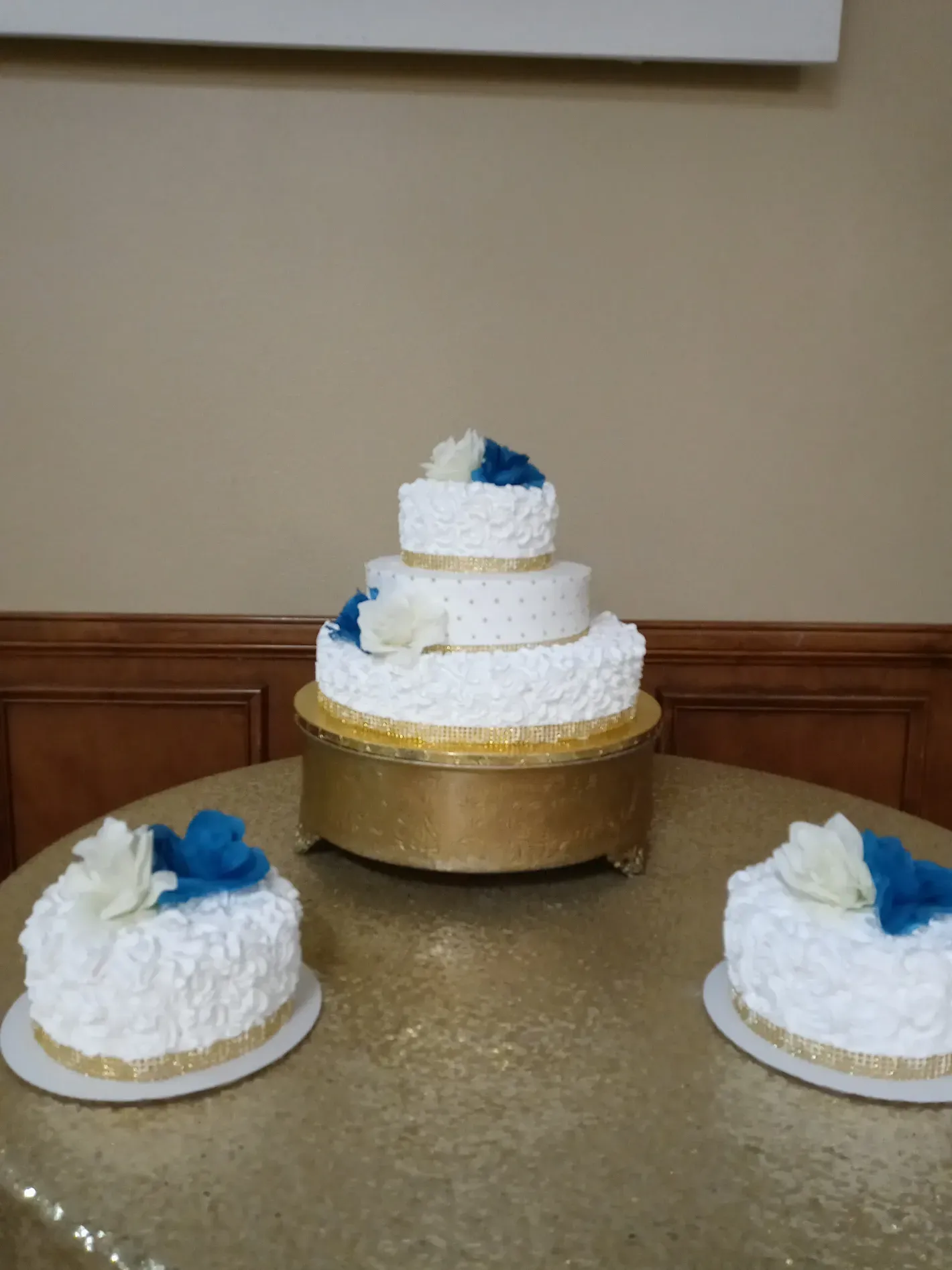 Three white tiered cakes with blue flowers on a gold tablecloth at a reception table