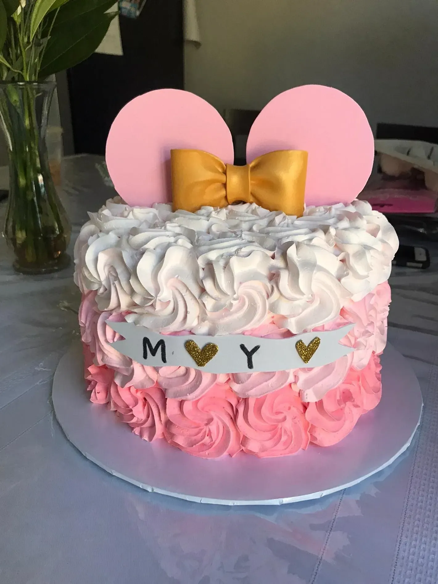 Mickey Mouse-themed pink and white frosted cake with gold bow and “M Y” banner on a stand