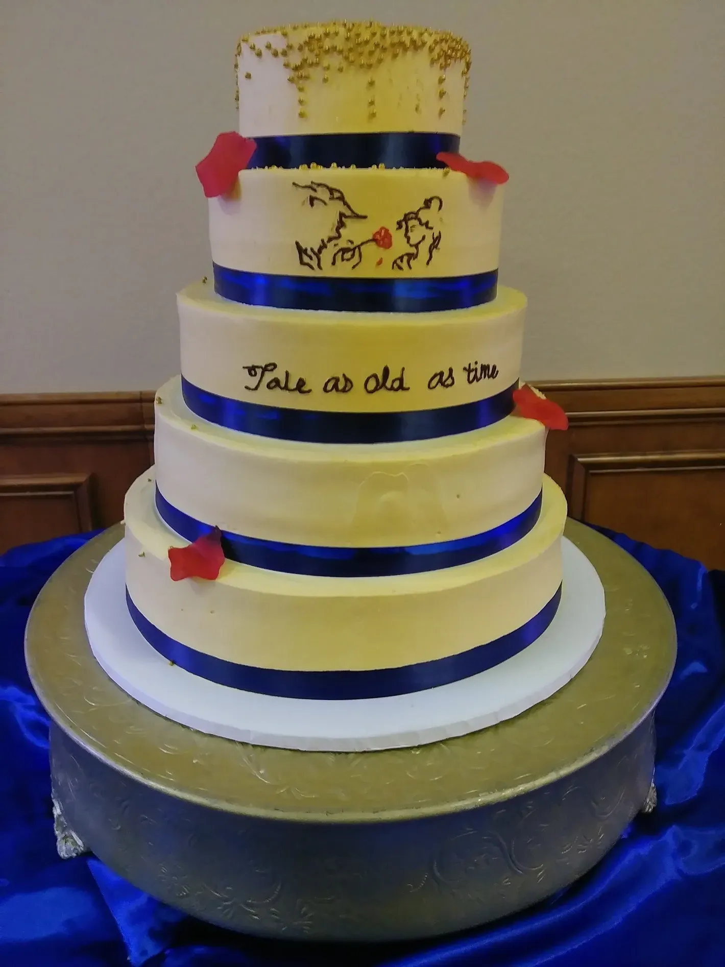 Five-tier white cake with blue ribbon bands and gold accents on a silver pedestal table