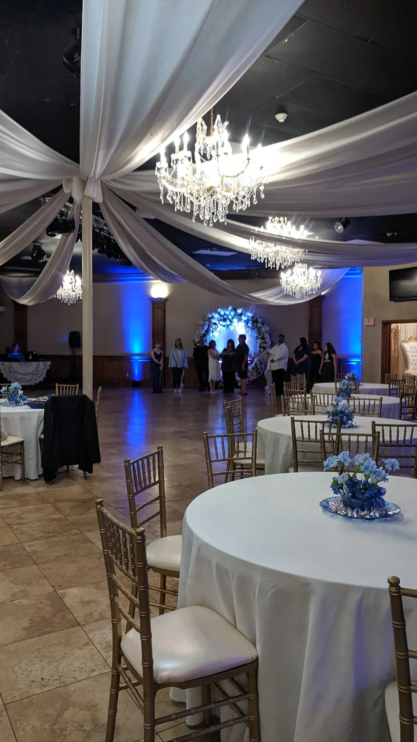 Elegant banquet hall with white draped ceiling, chandelier, round tables, and blue uplighting