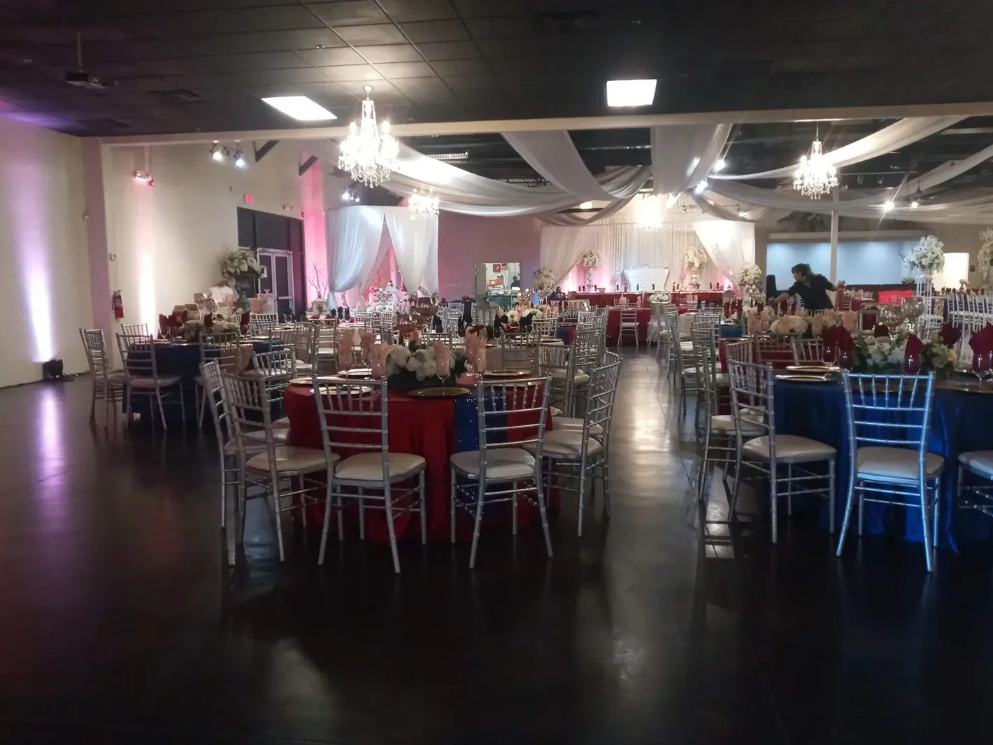 Banquet hall with round tables, red and blue chairs, and soft pink lighting under chandeliers