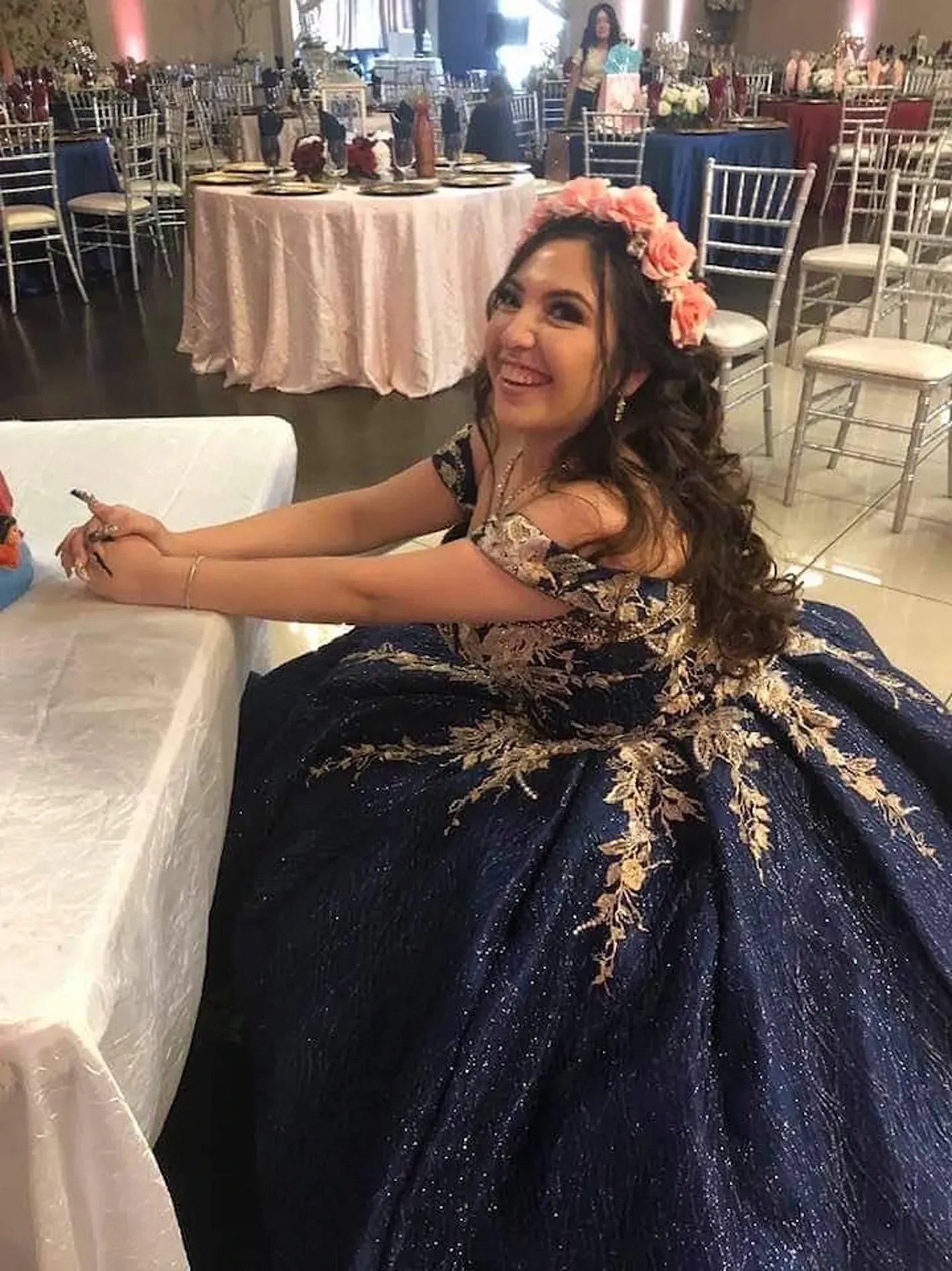 Woman in a navy gown with gold accents sitting at a decorated event table, smiling and writing.