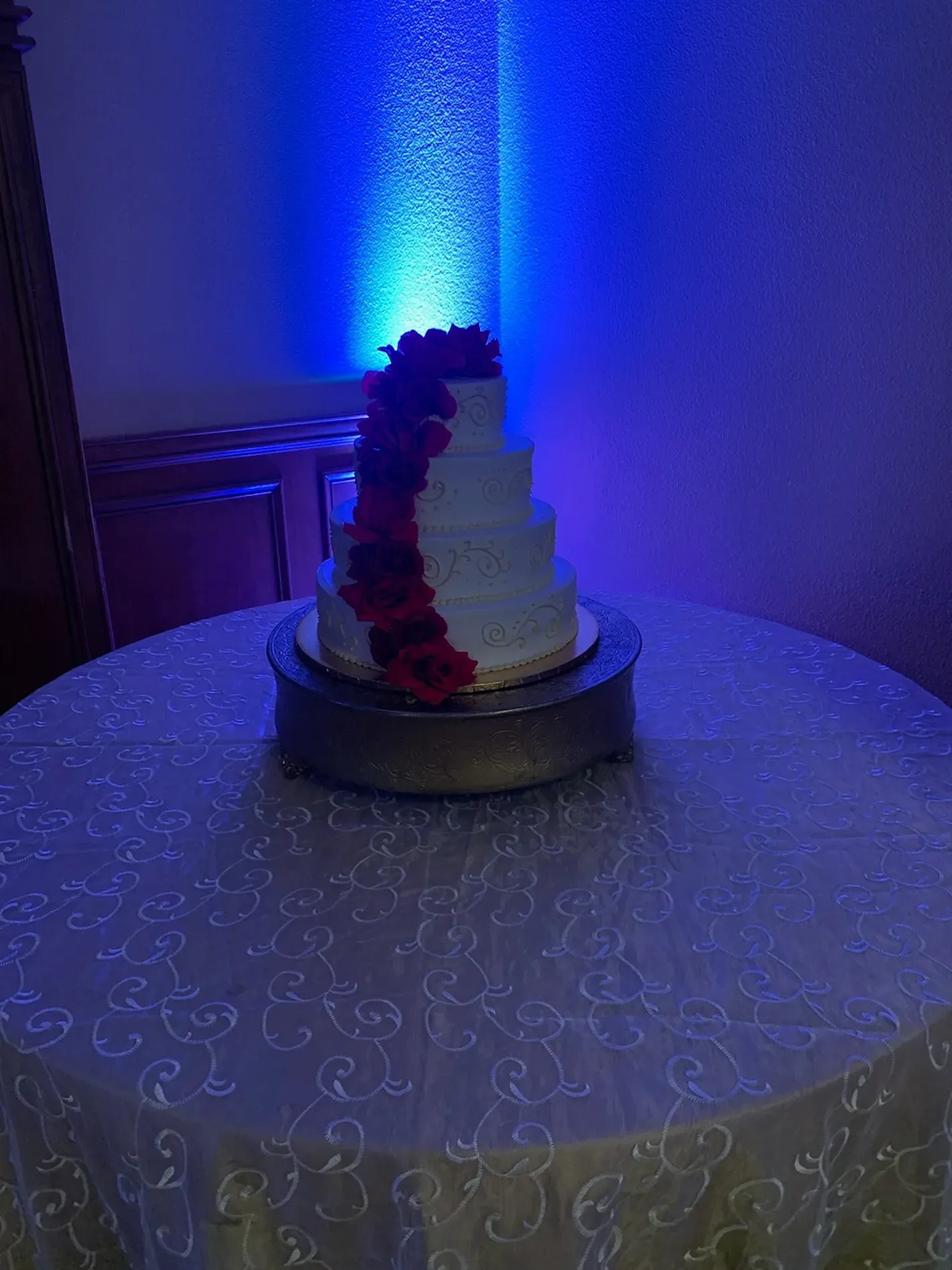 Tiered white cake with red and black roses on a round table, lit by blue ambient light.