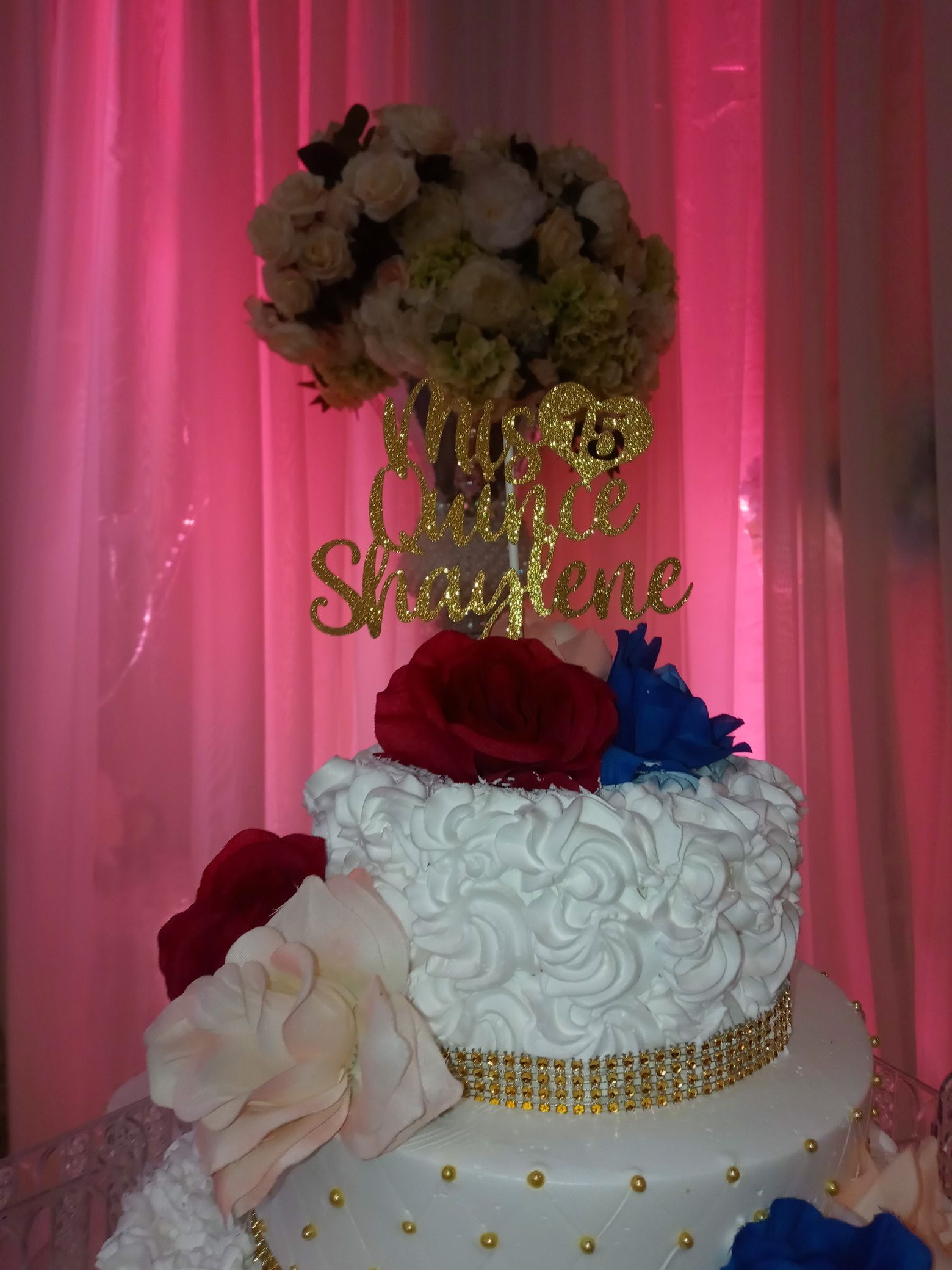 Two-tier white cake with red, blue, and pink flowers, gold accents, and “Sabrina” topper against pink drapes