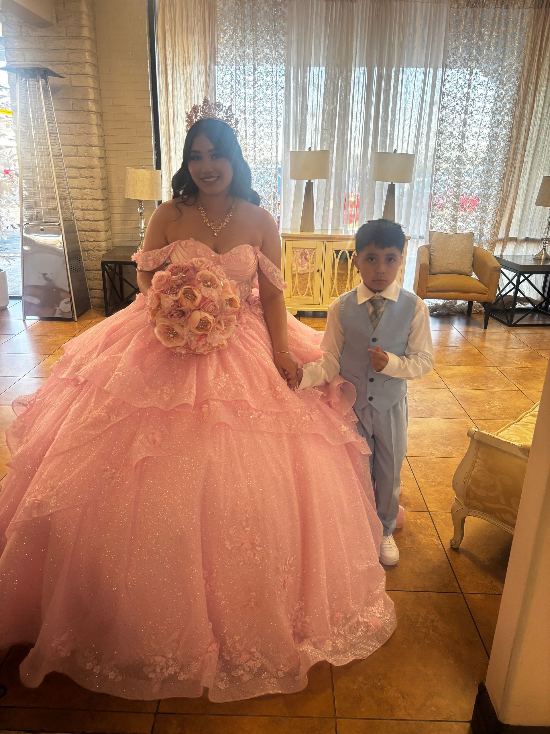 Person in a pink ball gown stands beside a child in a light suit in a bright room.