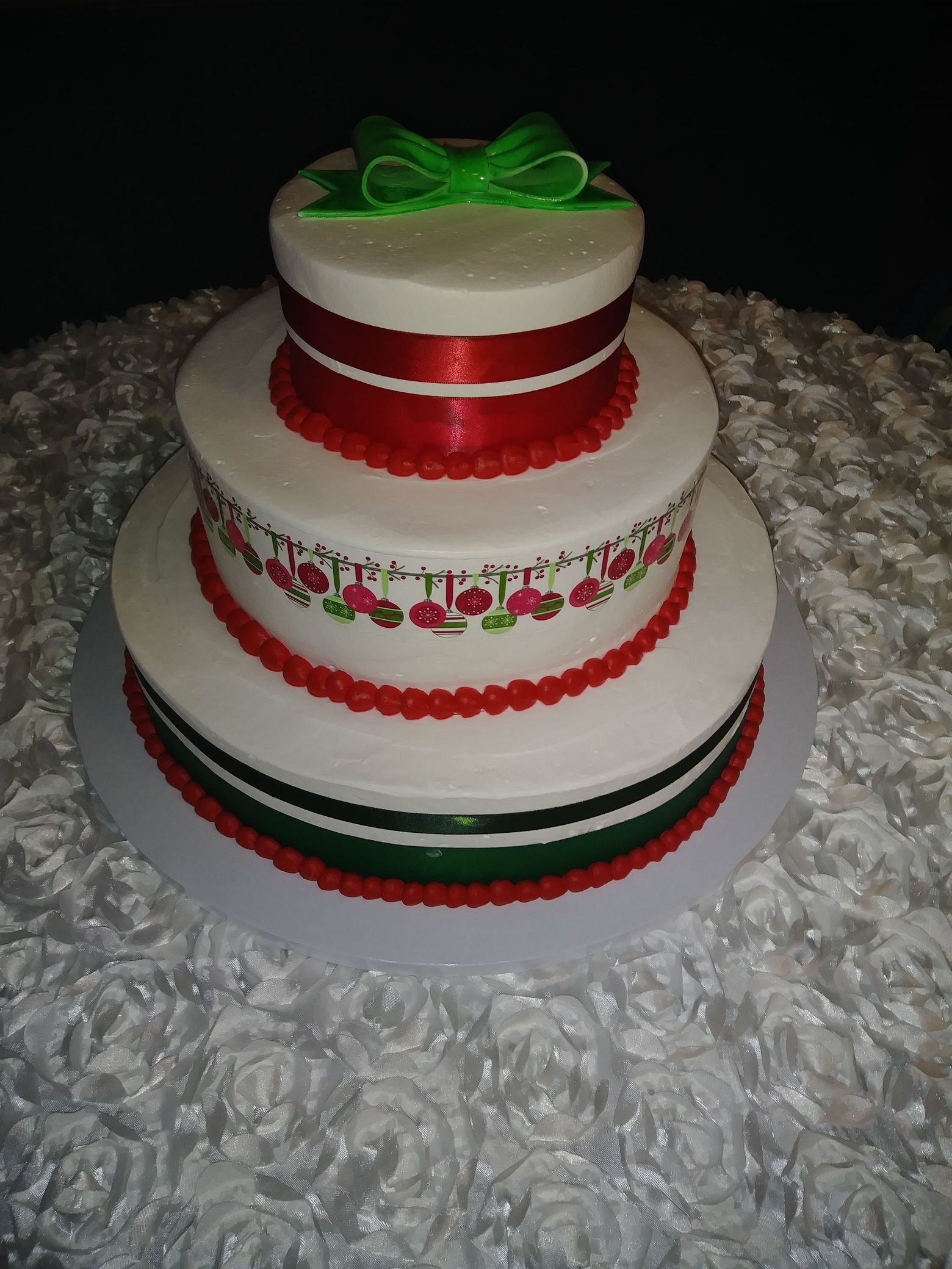Three-tier white cake with red ribbon bands and green bow, sitting on a textured white surface