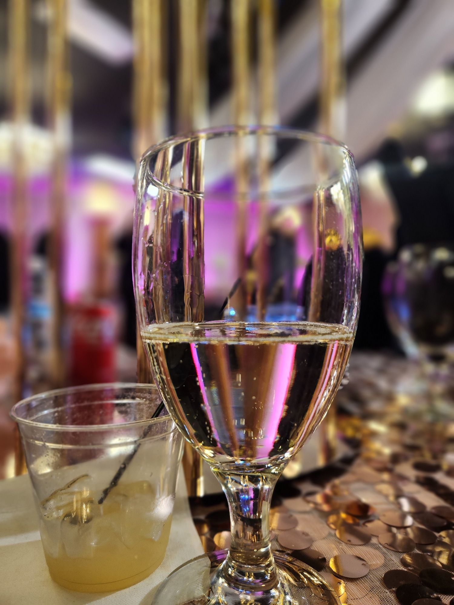 A wine glass and a plastic cup on a table, with pink-purple lights in a blurred background.