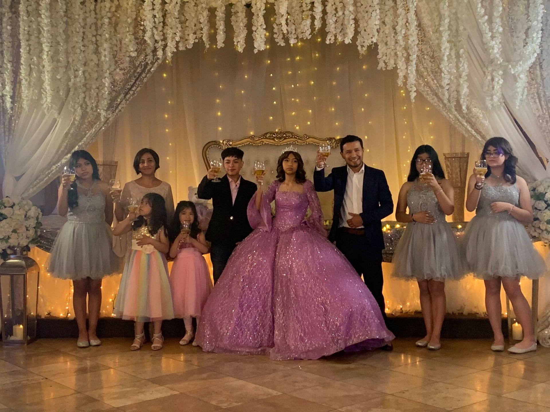 Seven people in formal attire pose in front of a sparkling draped backdrop, centered around a woman in a pink gown.