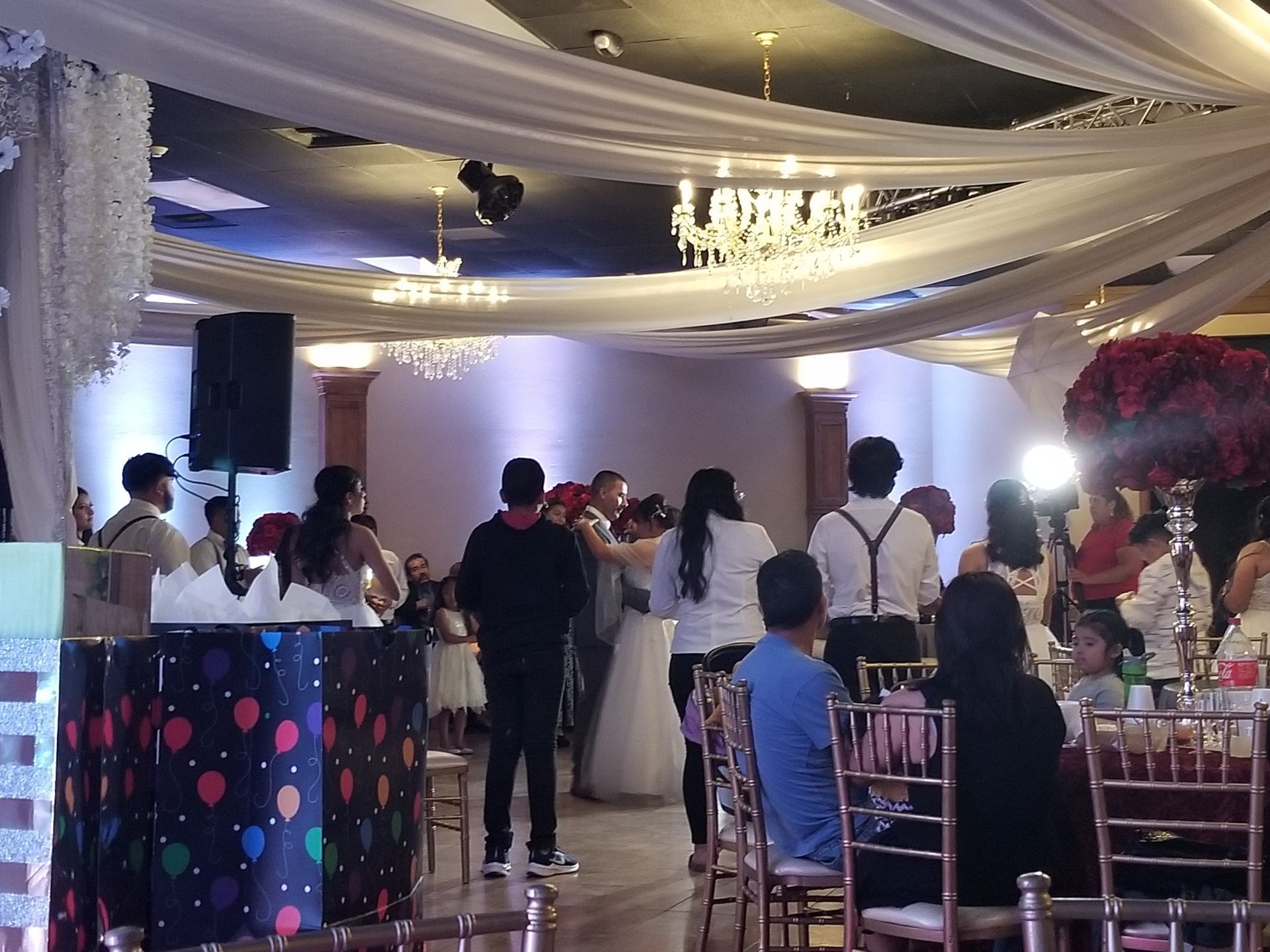 Indoor wedding reception with guests mingling under draped white and gold ceiling decor, chandeliers, and pink-lit tables