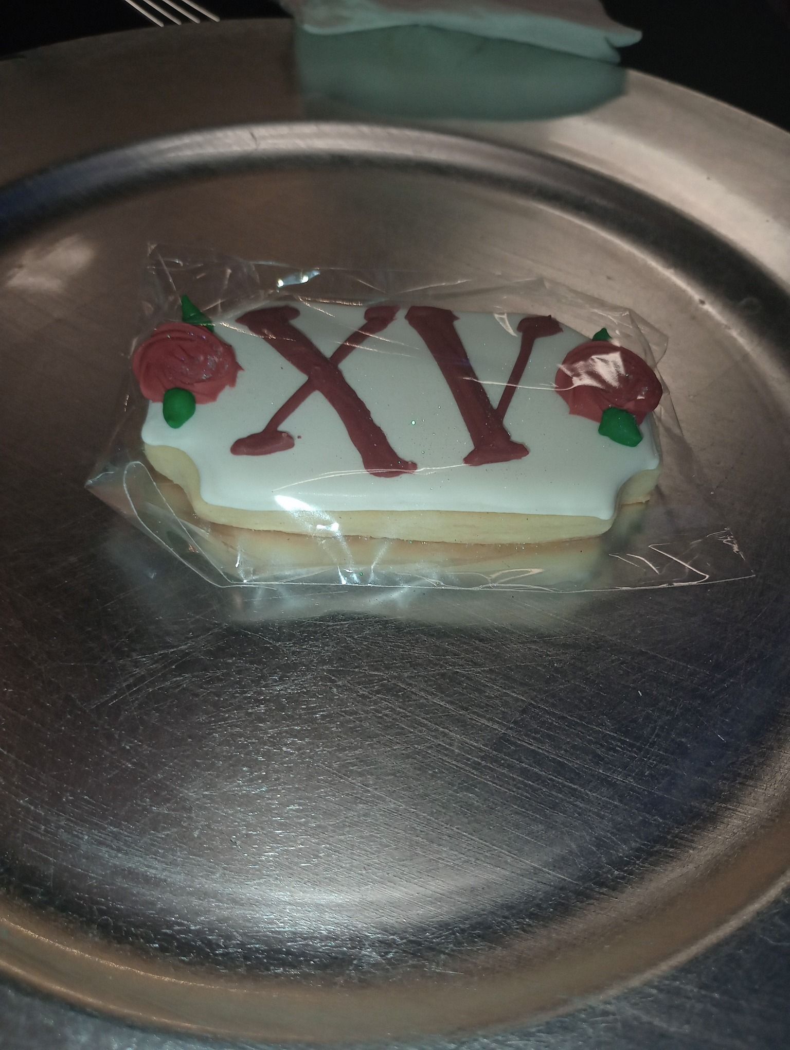 Dessert on a silver plate, topped with red “XV” and small strawberries, wrapped in plastic.