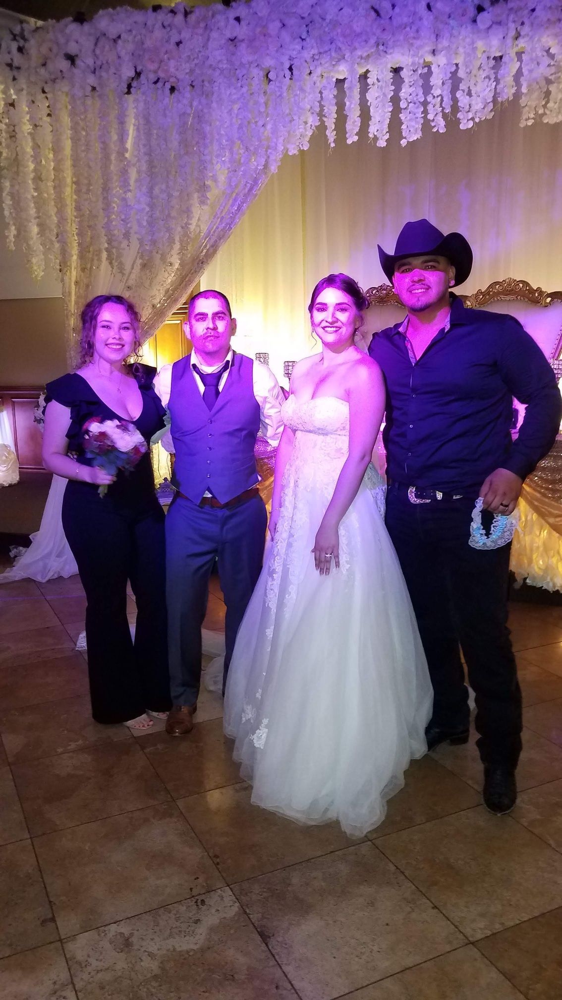 Four people posing together at a wedding reception, including the bride in a white dress and one man in a black cowboy hat.