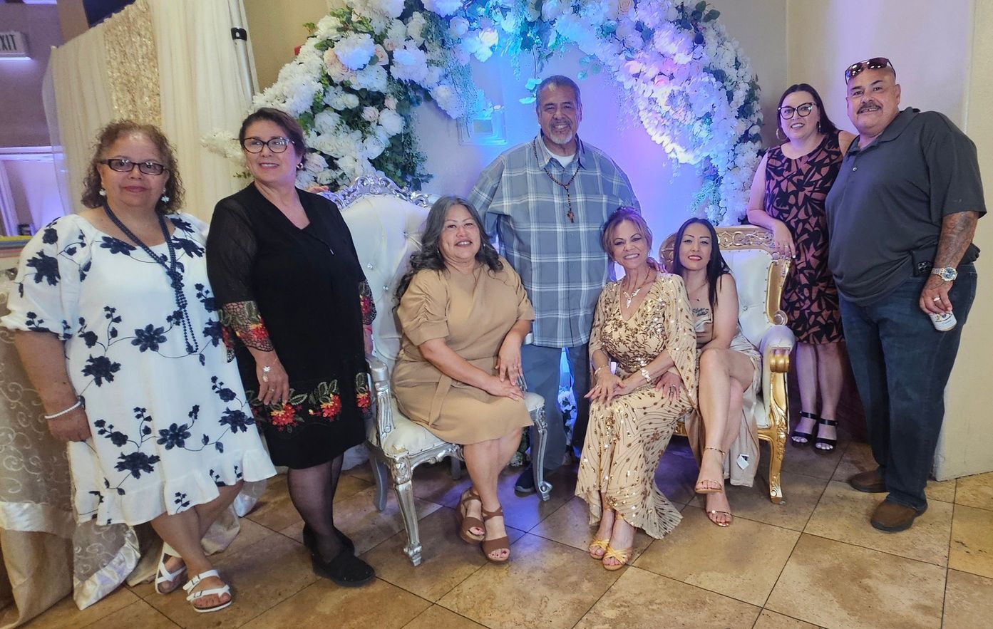 Group posing indoors under a floral arch with blue lights, several seated and standing, smiling at the camera.