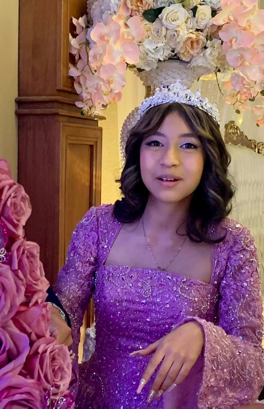 Woman in a purple sequined dress posing before a floral backdrop and wooden paneling.