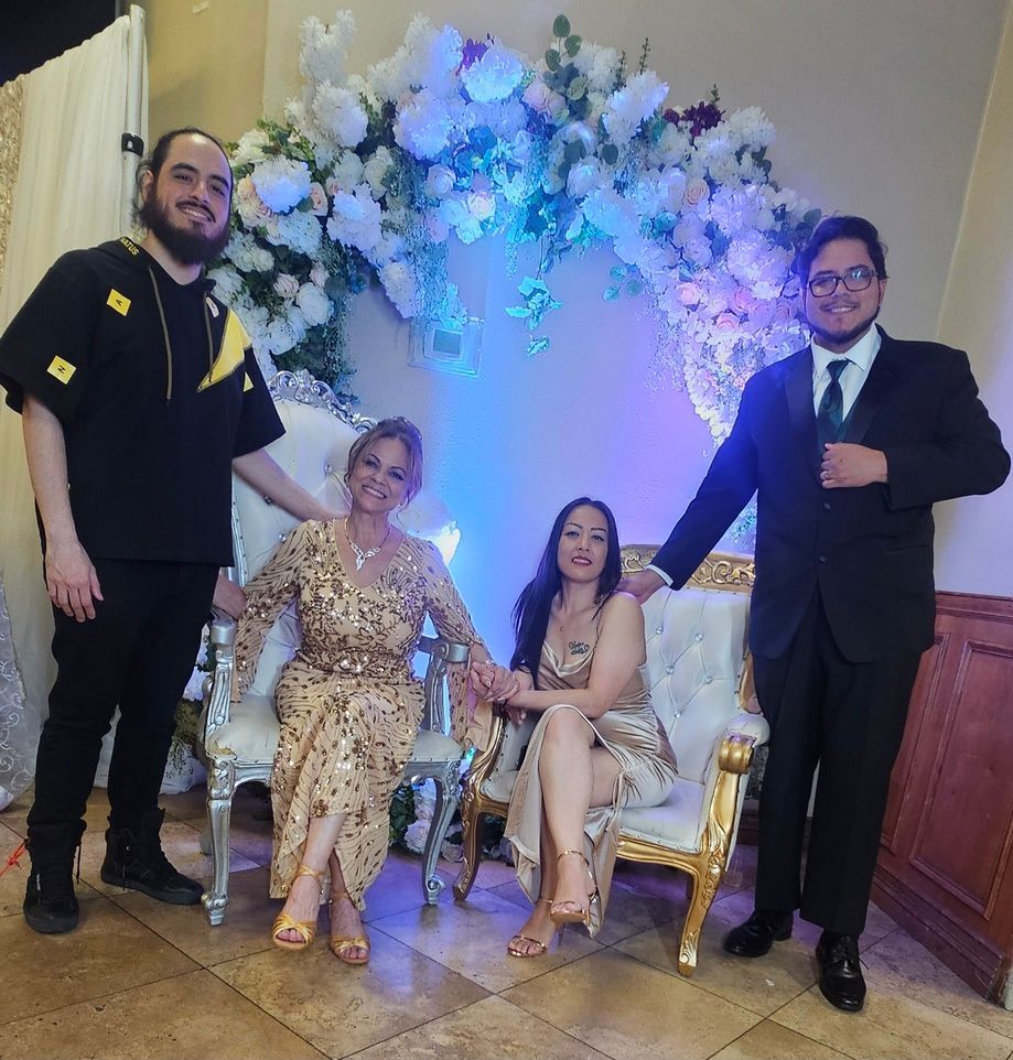 Four people posing at an event under a white-and-blue floral arch, two seated and two standing in formal attire