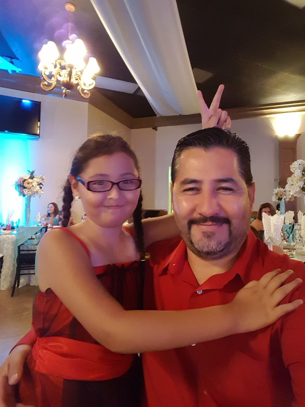 Two people in red outfits pose indoors at a celebration, smiling, with the woman making bunny ears behind the man's head
