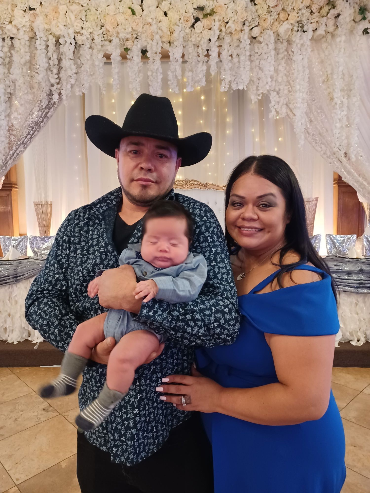 A couple holding a baby in front of a white floral backdrop, with the man wearing a black cowboy hat.