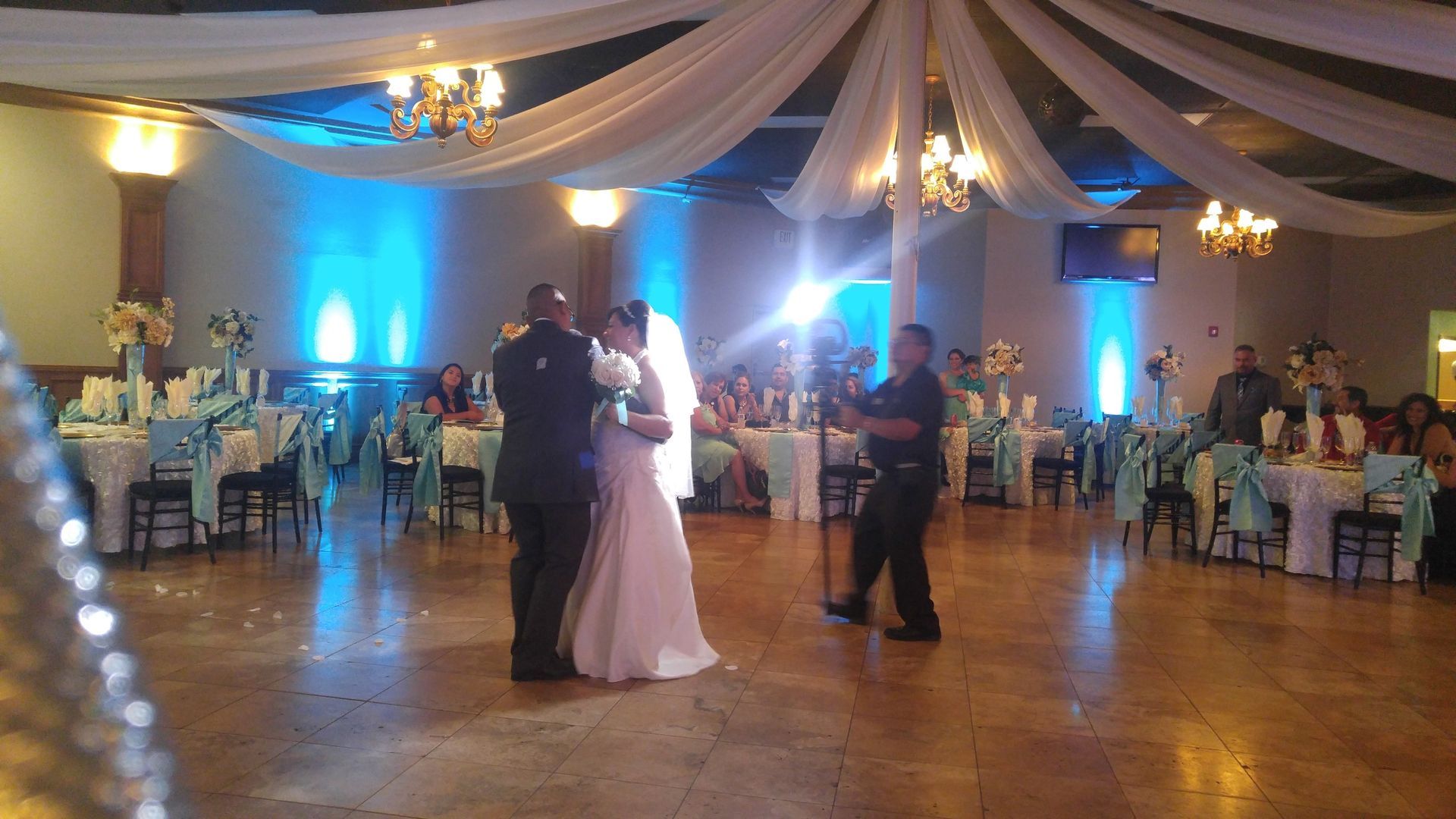 Bride and groom dancing at a reception hall with blue uplighting and seated guests nearby