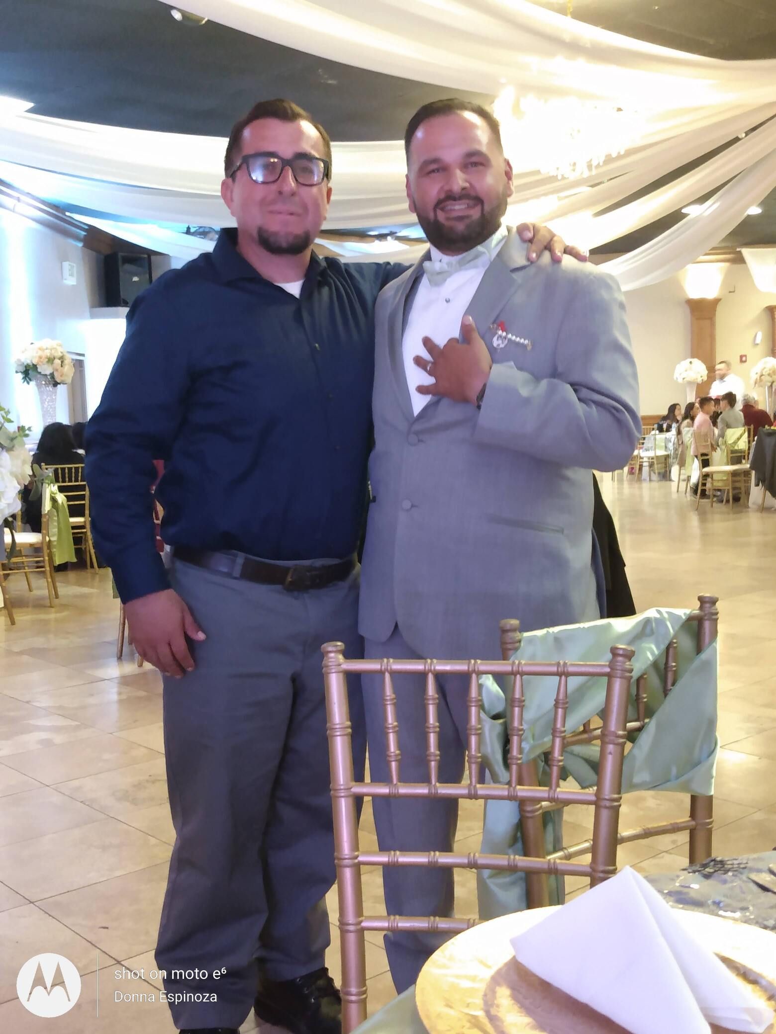 Two men pose together at a decorated indoor event, one in a light gray suit and one in a dark shirt.