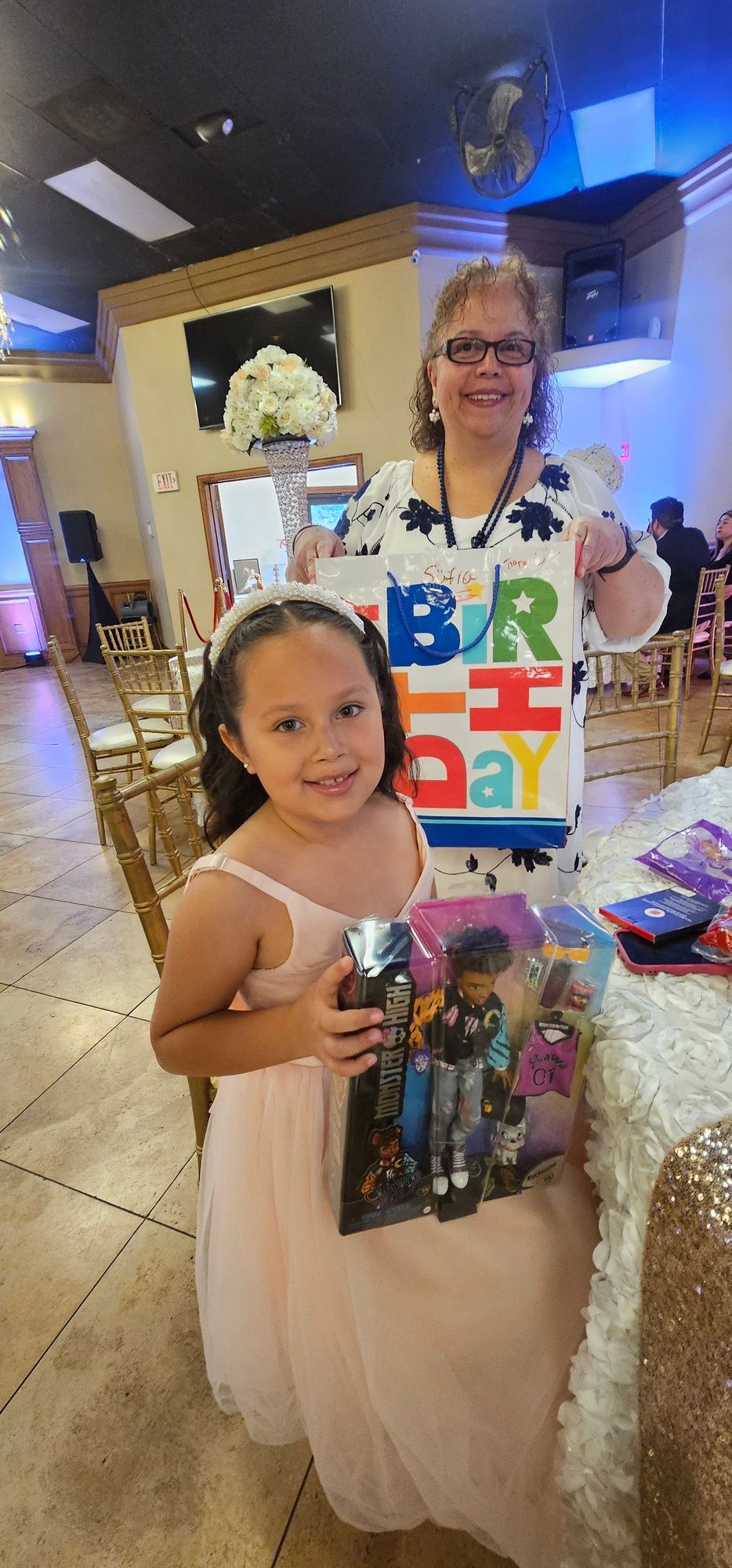 Child in a white dress holding a gift bag, standing with an adult in a decorated indoor party setting