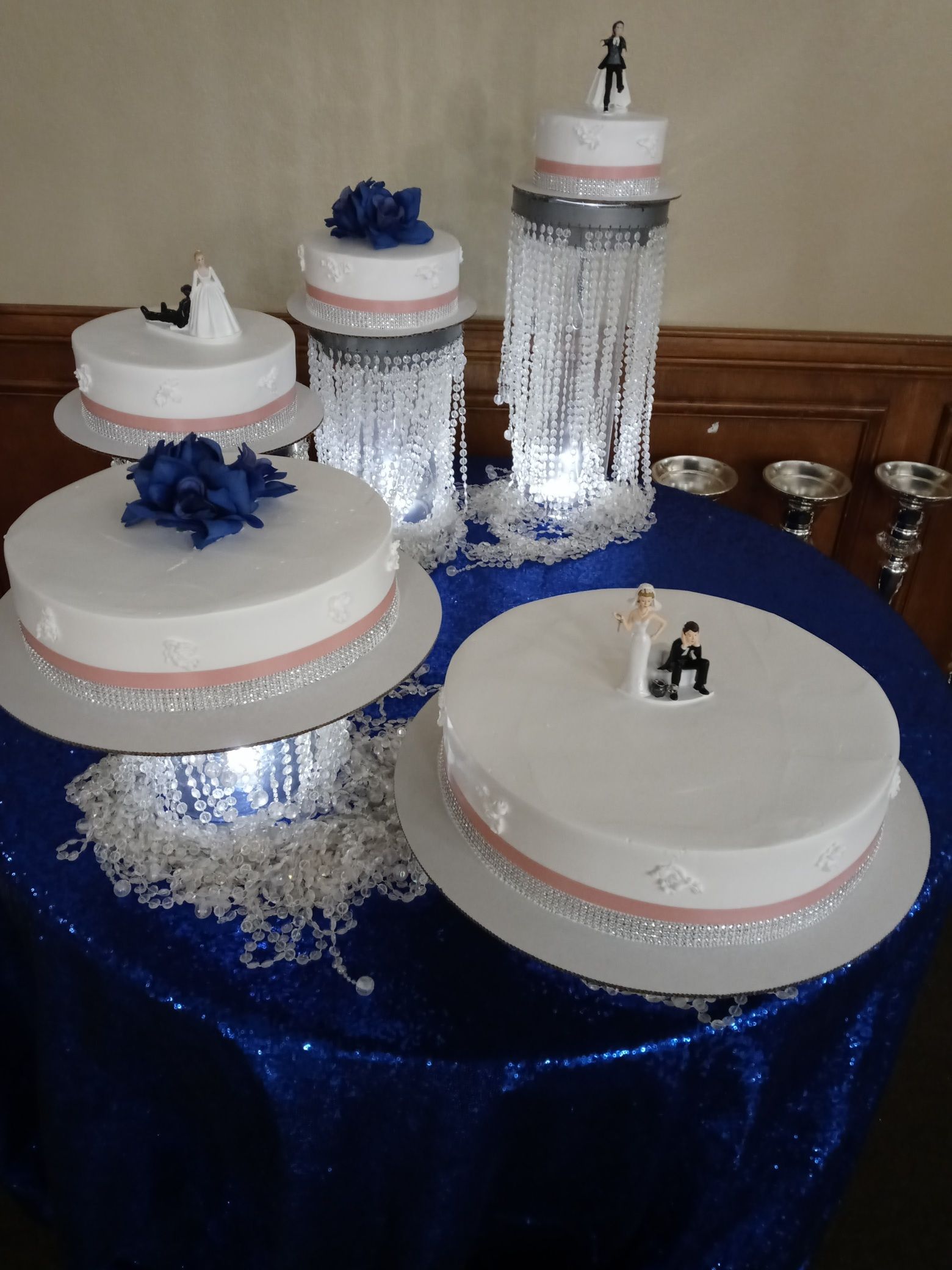 White tiered cakes with blue accents displayed on a royal blue tablecloth at an event table