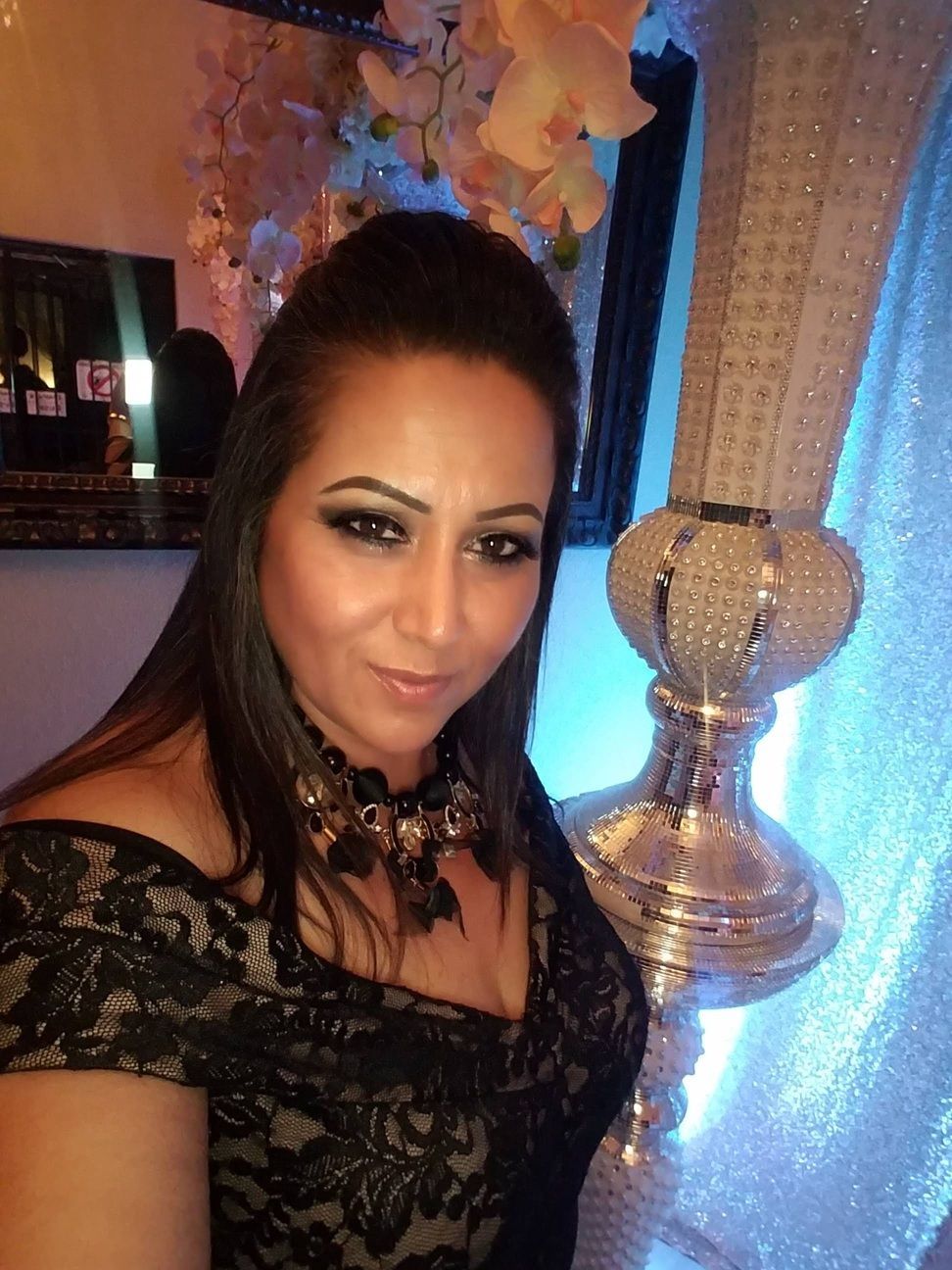 Woman in a black lace dress posing indoors beside a decorative silver lamp.